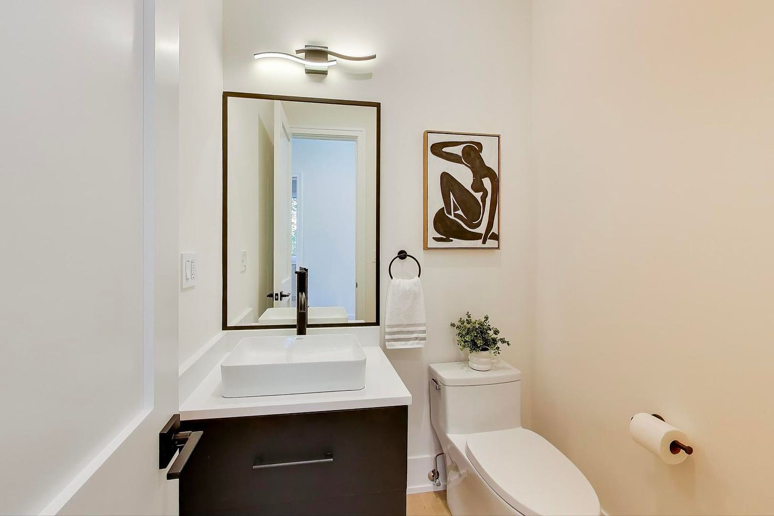 This is a well-lit guest bathroom featuring a modern design. The room includes a floating vanity with a white countertop and a square vessel sink, complemented by a black faucet and a dark wood cabinet. A framed mirror hangs above the sink, and a piece of art adorns the wall above the toilet, which is equipped with a bidet.