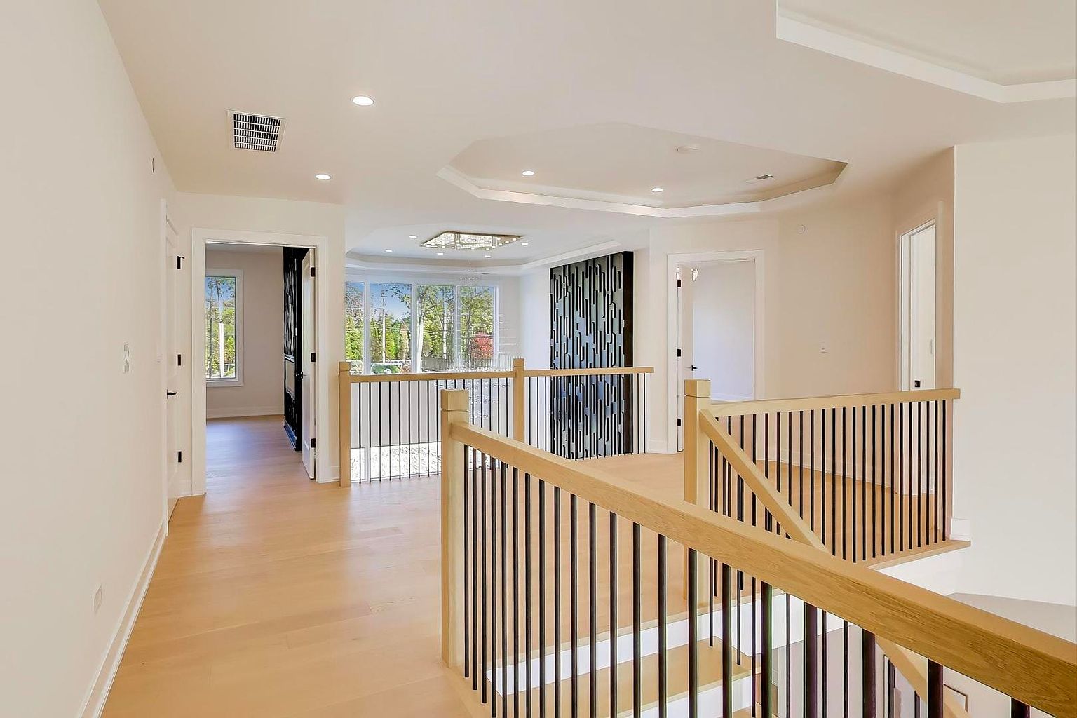 This is an interior shot of a bright and airy hallway and staircase area. The space features light wood flooring, white walls, and a modern railing system with light wood handrails and black metal spindles. The hallway leads to other rooms, visible through doorways, and the staircase descends to another level, creating a sense of openness and flow.