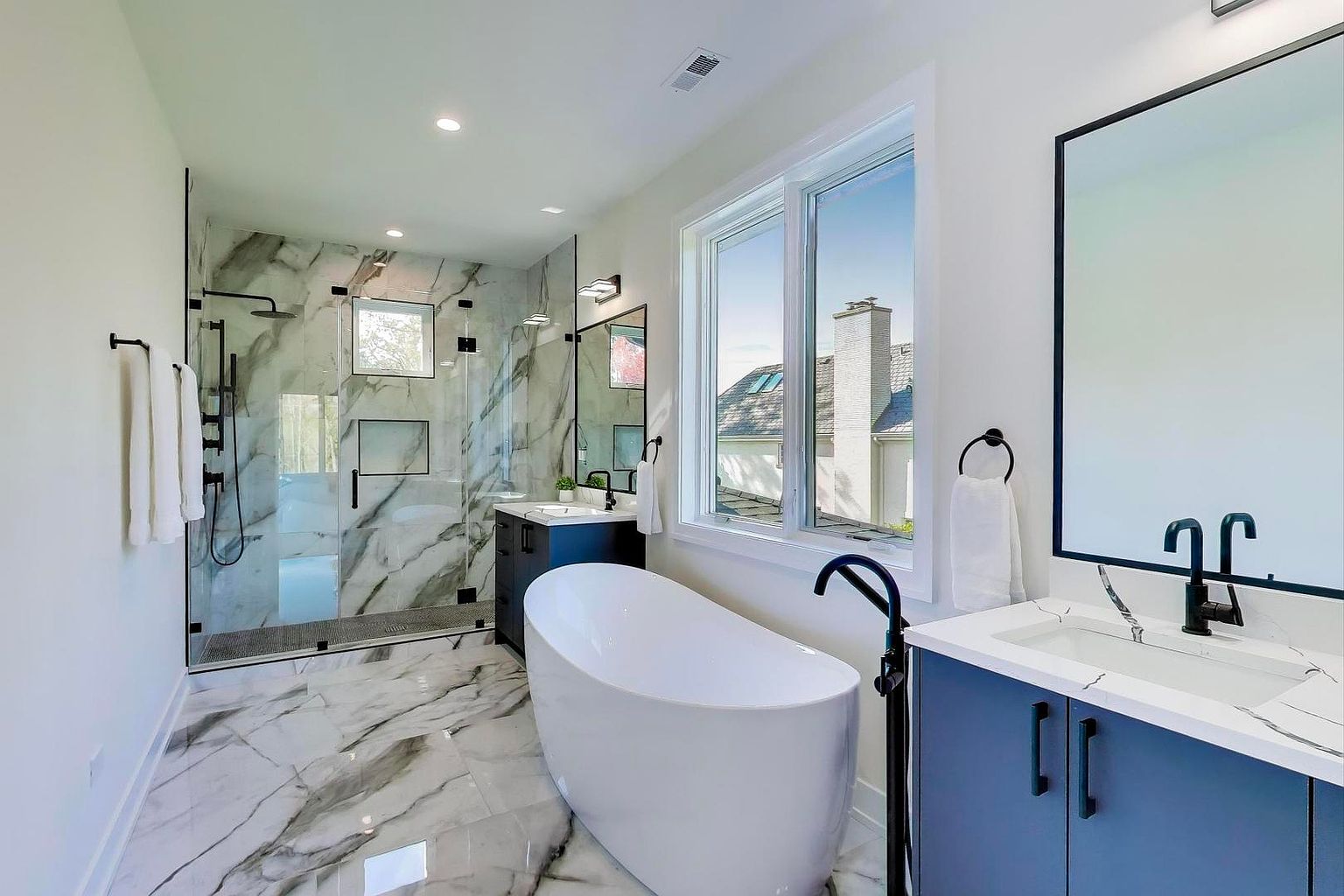 This is a modern primary bathroom featuring marble flooring and a glass-enclosed shower with black hardware. A freestanding white bathtub sits next to a large window, providing natural light. The vanity is a dark blue with a white countertop and black fixtures, creating a sleek and luxurious feel.