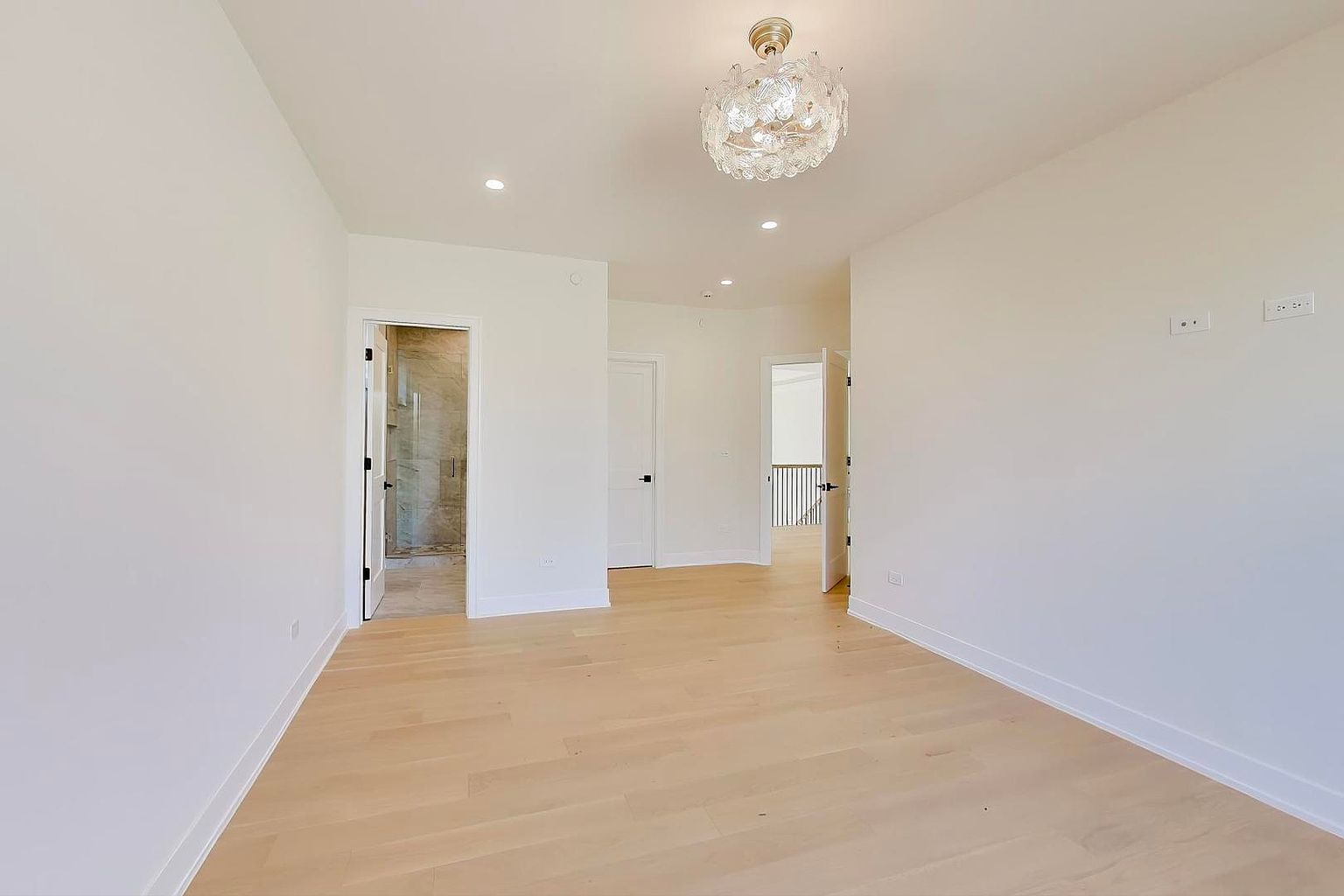 This is a bright and spacious primary bedroom featuring light hardwood floors and white walls. The room has multiple doorways leading to other areas, including a bathroom and hallway. A decorative chandelier hangs from the ceiling, adding a touch of elegance to the minimalist design.
