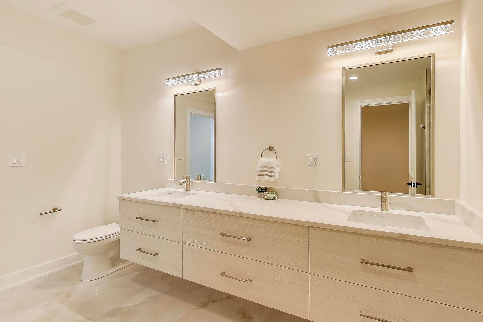 This is a bright and modern primary bathroom featuring a double vanity with light wood-grain cabinets and a white countertop. Two large mirrors are illuminated by contemporary light fixtures, and a toilet is visible to the left. The flooring appears to be marble or a marble-like material, adding to the luxurious feel of the space.