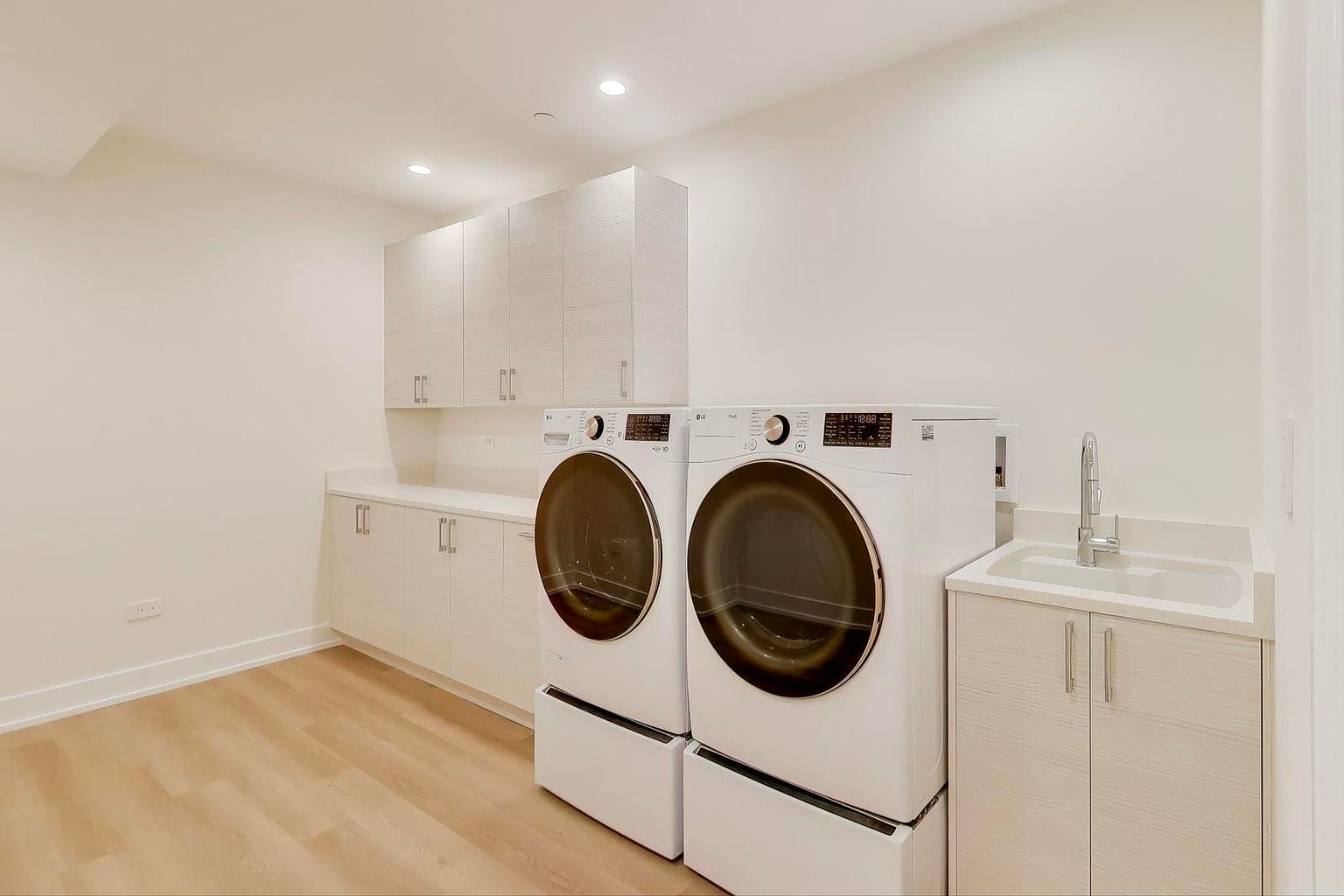 This is a well-lit laundry room featuring a modern design with light wood-look flooring and white walls. The room is equipped with a front-loading washer and dryer set, both elevated on pedestals, and a sink with a chrome faucet set into a cabinet. Built-in cabinets above the countertop provide ample storage space, creating a clean and organized area.
