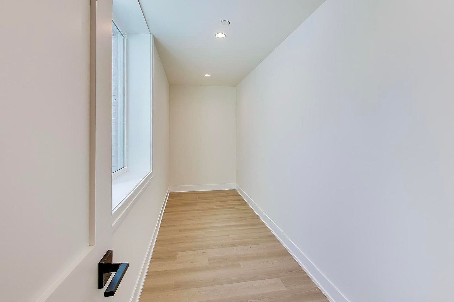 This is a well-lit hallway with light wood flooring and white walls, creating a clean and modern aesthetic. The hallway features a window on the left side, providing natural light, and recessed lighting in the ceiling. The perspective is from one end of the hallway, emphasizing its length and minimalist design.