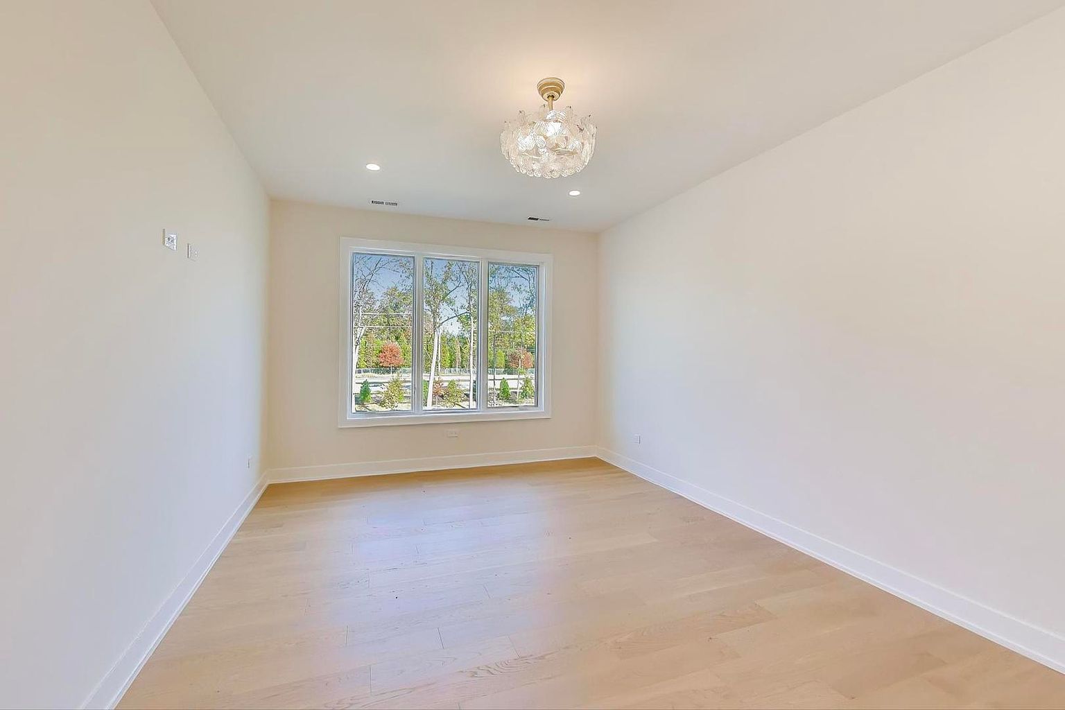 This is a bright and airy bedroom featuring light hardwood floors and white walls, creating a clean and modern aesthetic. A large window provides ample natural light and a view of the outdoors. The room is illuminated by a decorative chandelier, enhancing the space's elegance and appeal.