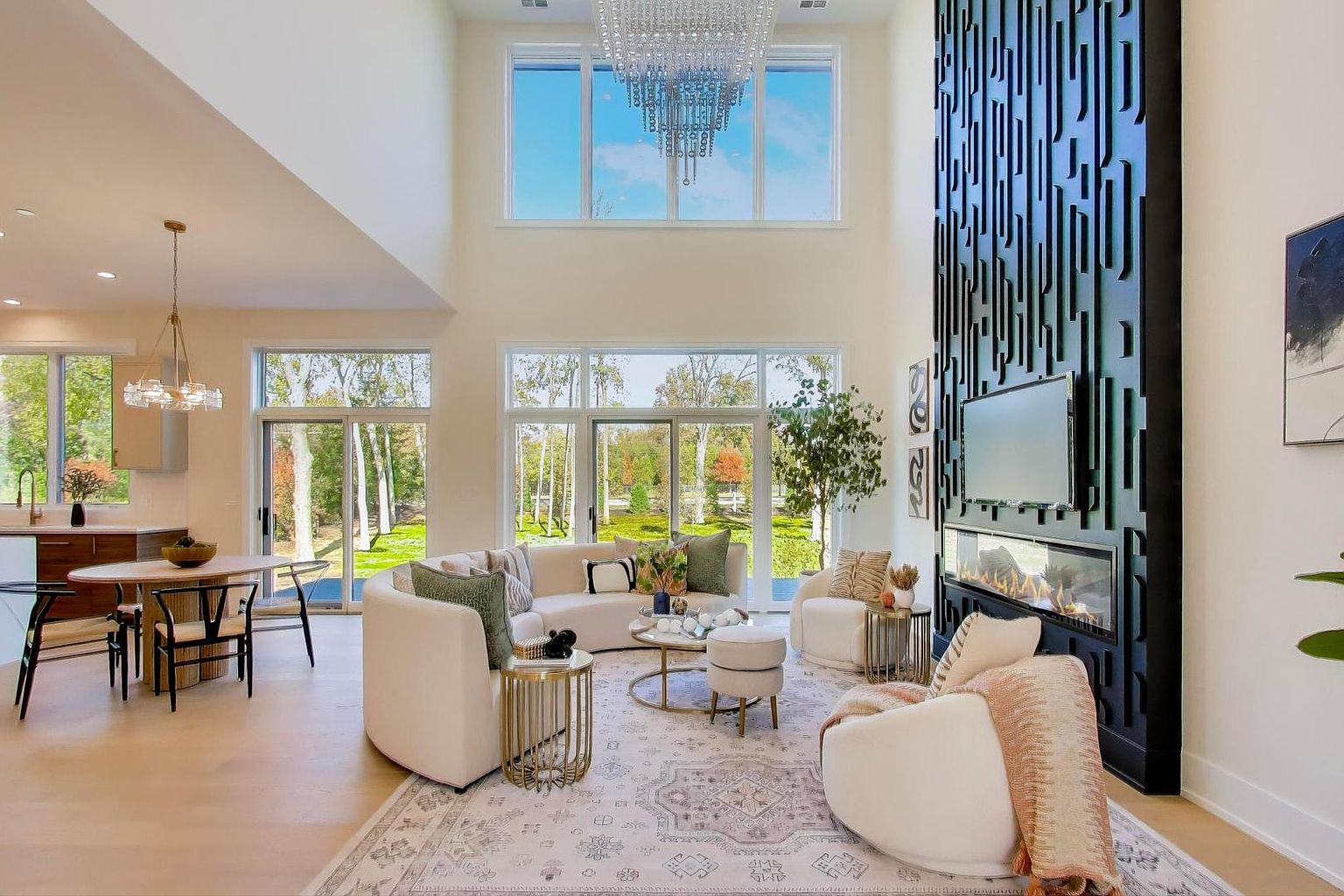 This is a spacious and luxurious living room featuring high ceilings and large windows that provide ample natural light. The room is furnished with a curved sofa, armchairs, a round coffee table, and a modern fireplace with a decorative wall feature. The overall style is contemporary and elegant, creating a welcoming and sophisticated atmosphere.