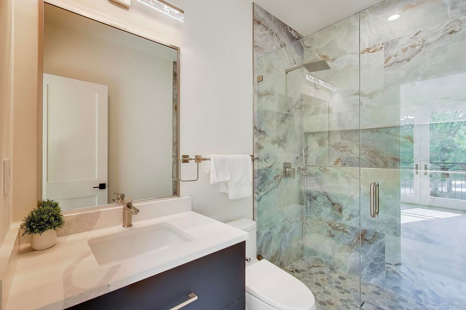 This is a modern bathroom featuring a vanity with a white countertop and dark blue cabinetry, a large mirror, and a glass-enclosed shower with marble-like tiling. The shower has a rainfall showerhead and a pebble floor. The overall impression is clean, stylish, and luxurious.