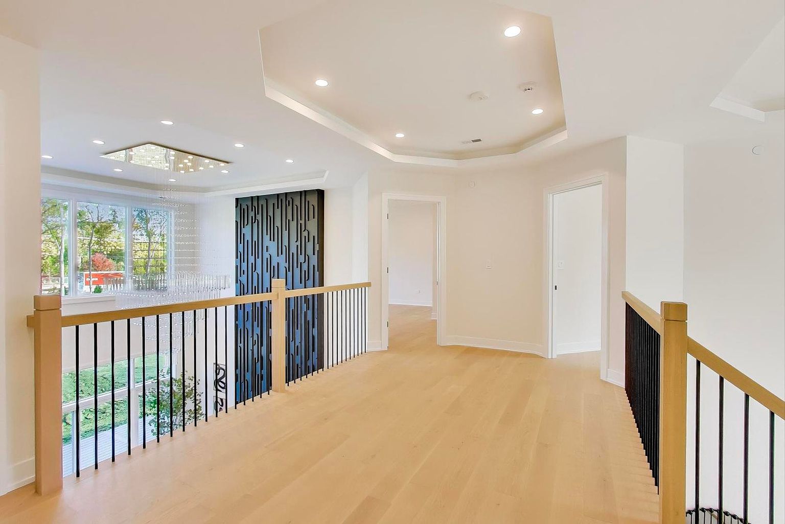 This is an interior shot of a hallway on an upper level, featuring light hardwood flooring and a modern railing with wooden posts and black metal spindles. The hallway leads to several doorways, and there's a decorative black panel on the left. The space is well-lit with recessed lighting, and the overall impression is clean and contemporary.