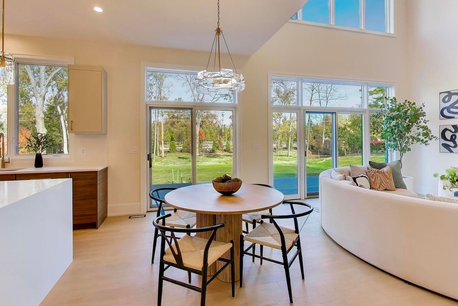 This is an interior shot of a dining area adjacent to a living space, featuring a round wooden table with four chairs. Large windows provide ample natural light and views of the outdoor greenery. The space is modern and airy, with a neutral color palette and minimalist decor, creating an inviting and open atmosphere.