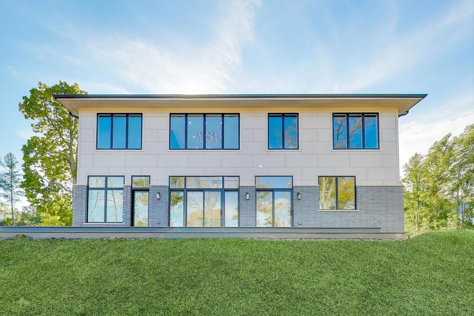 This is a front exterior view of a modern two-story house. The house features a combination of light-colored siding on the upper level and gray brick on the lower level, with large windows framed in black. A well-maintained green lawn slopes gently towards the house, creating a welcoming and upscale impression.