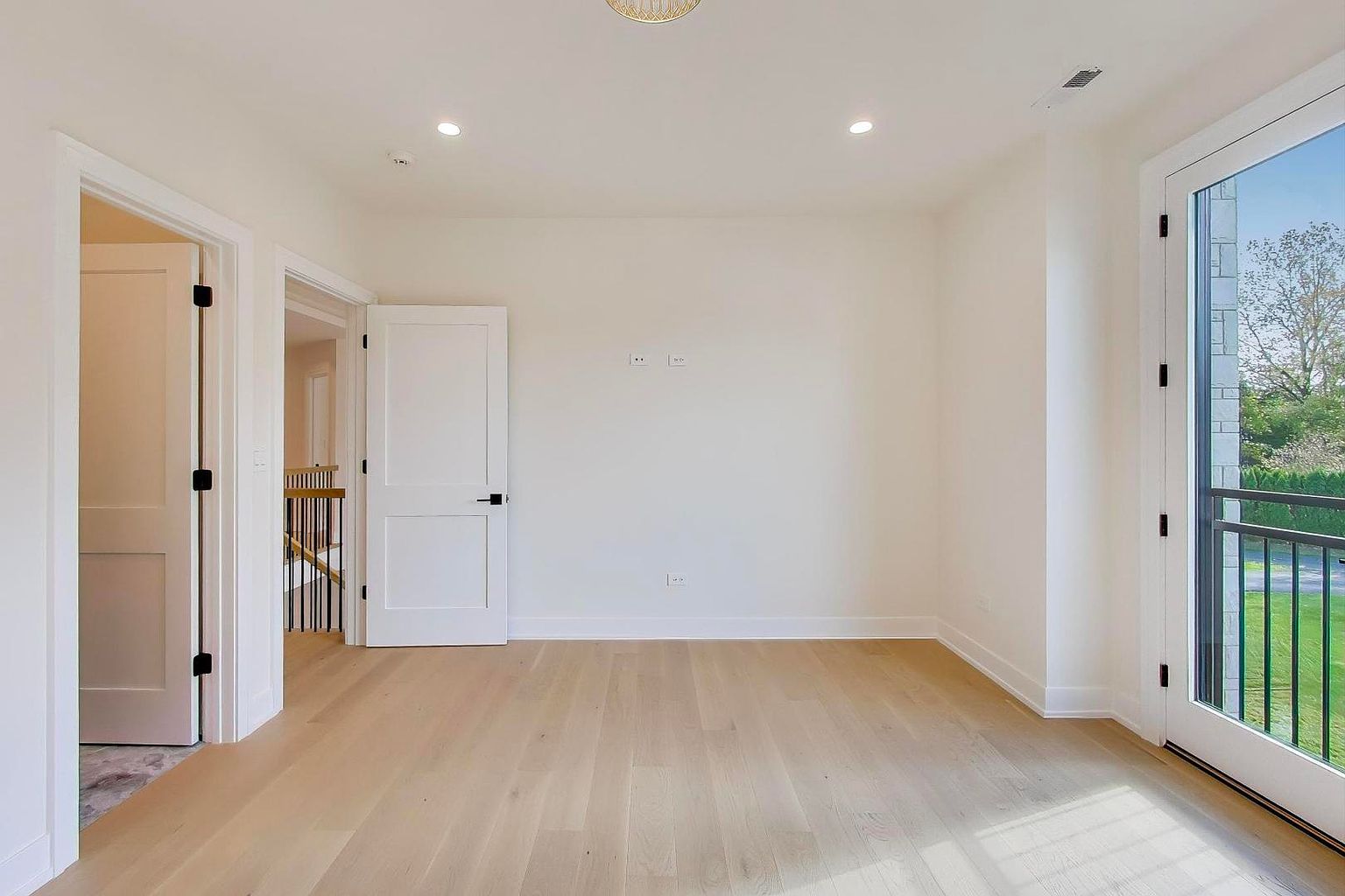This is a bright and airy bedroom featuring light hardwood floors and neutral-colored walls. The room has multiple doorways and a glass door leading to a balcony, providing ample natural light. The overall impression is clean and modern, creating a welcoming space.