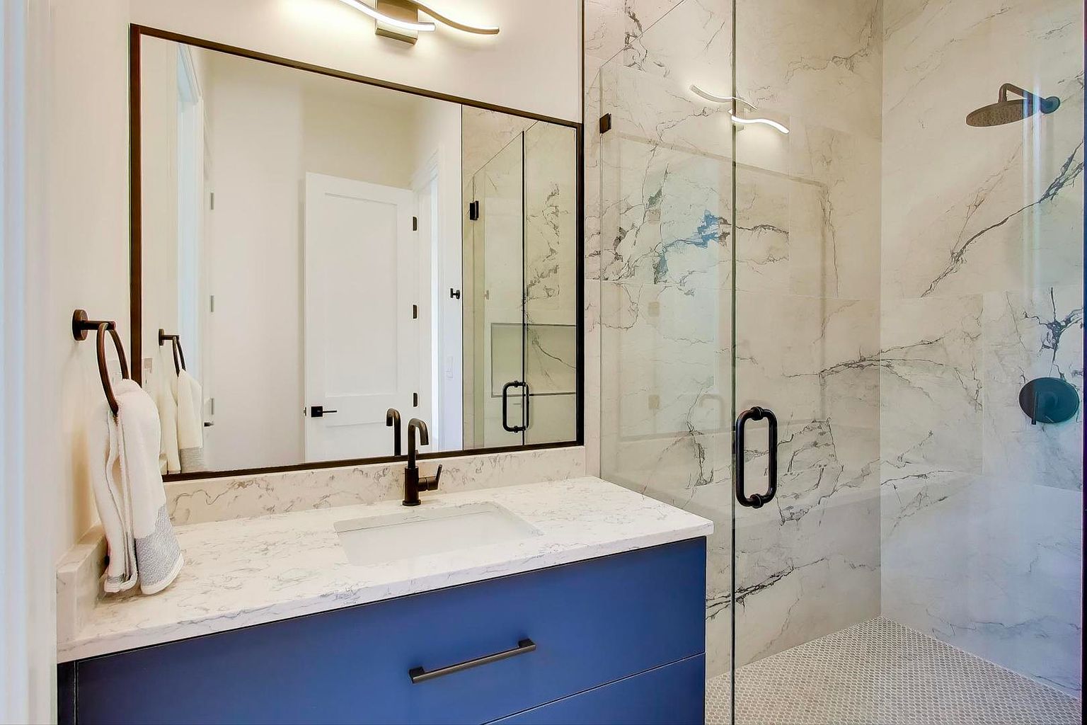 This is a modern primary bathroom featuring a blue vanity with a white countertop and a rectangular sink. A large mirror with a dark frame hangs above the vanity, reflecting a white door. The shower is enclosed with glass and has marble-like tile, creating a luxurious and clean aesthetic.
