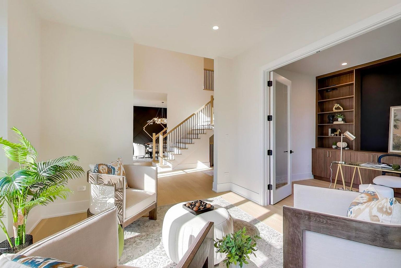 This interior shot showcases a bright and airy living room with a staircase in the background. The room features neutral-toned armchairs, a white ottoman, and a view into an adjacent office space through a glass door. Natural light floods the space, highlighting the hardwood floors and creating a welcoming atmosphere.