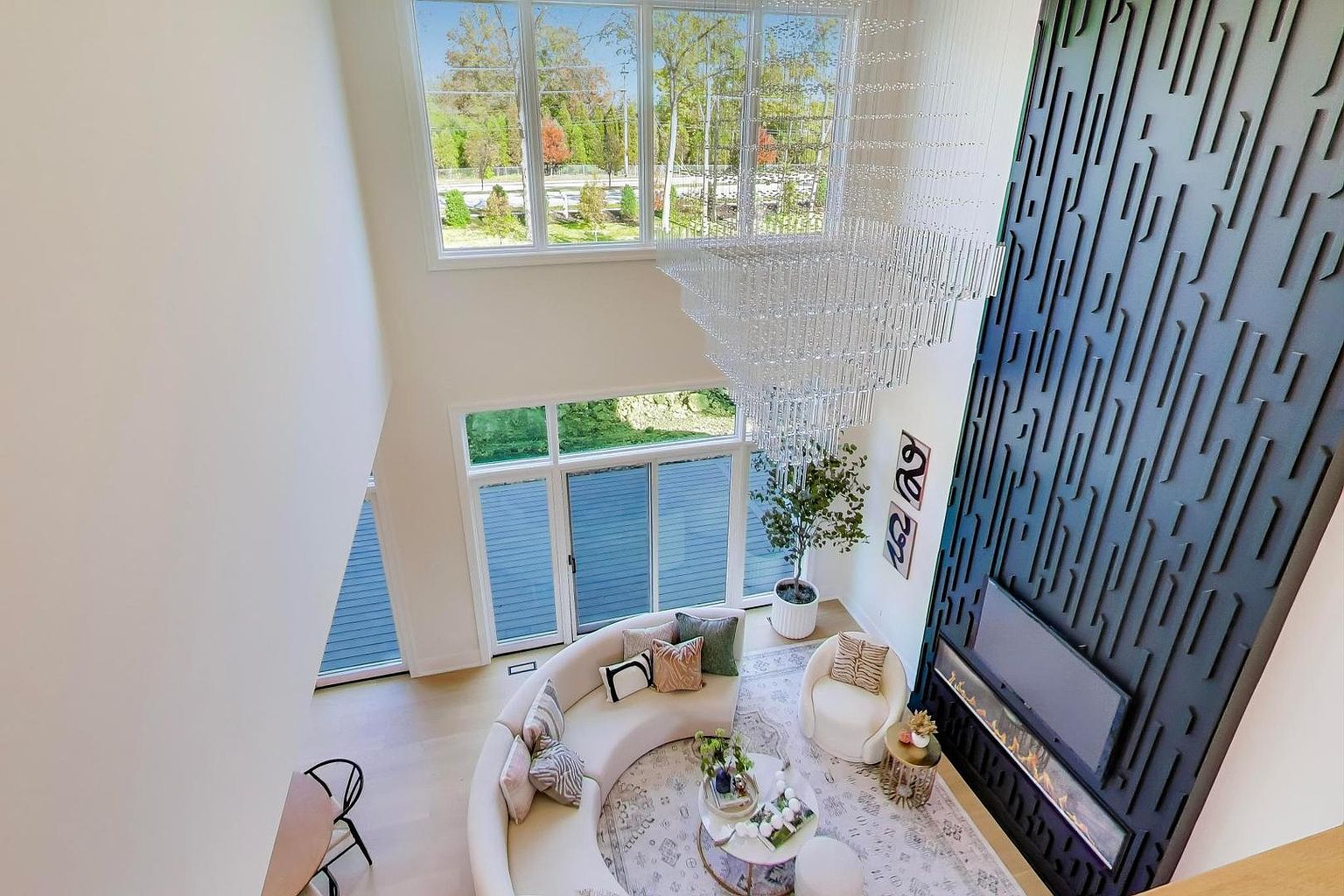 This is a high-angle shot of a luxurious living room featuring a curved sofa, a modern fireplace with a decorative black wall, and a large crystal chandelier. The room is bright and airy, with large windows offering natural light and views of the outdoors. The overall impression is one of elegance and sophistication.