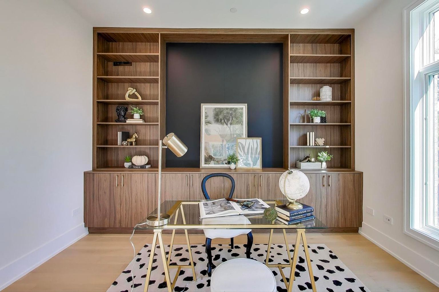 This is an interior shot of a home office or study, featuring a built-in wooden bookcase with open shelving and closed cabinets. A glass-topped desk with gold legs sits in the center of the room, complemented by a black and white patterned rug and a chair. The room is well-lit, creating a sophisticated and functional workspace.
