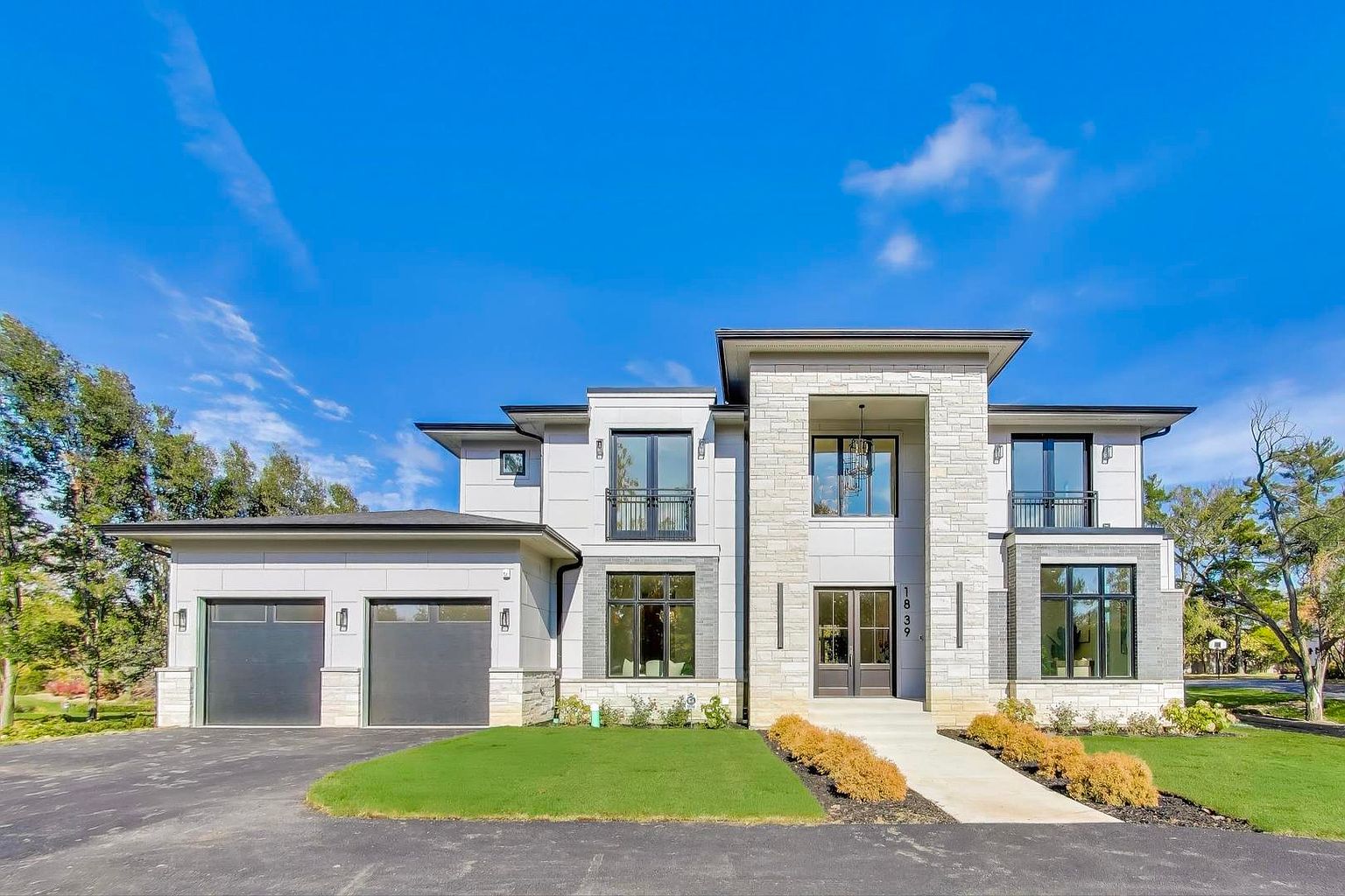 This is a front view of a modern, two-story house with a prominent stone facade and a two-car garage. The house features large windows, some with balconies, and a well-manicured lawn. The driveway is paved and leads to the garage, enhancing the property's curb appeal.