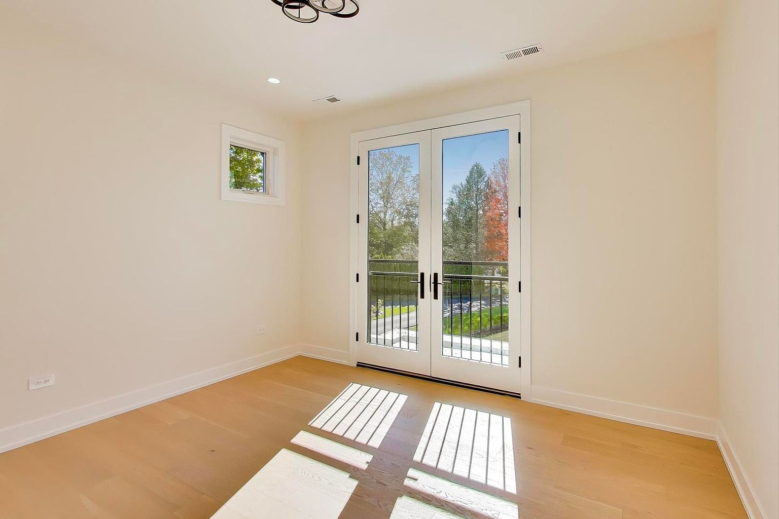 This is a bright and airy bedroom featuring light hardwood floors and neutral-colored walls. A set of French doors leads to a balcony, providing ample natural light and a view of the outdoors. A small window on the left wall adds to the room's brightness, creating a welcoming and serene atmosphere.