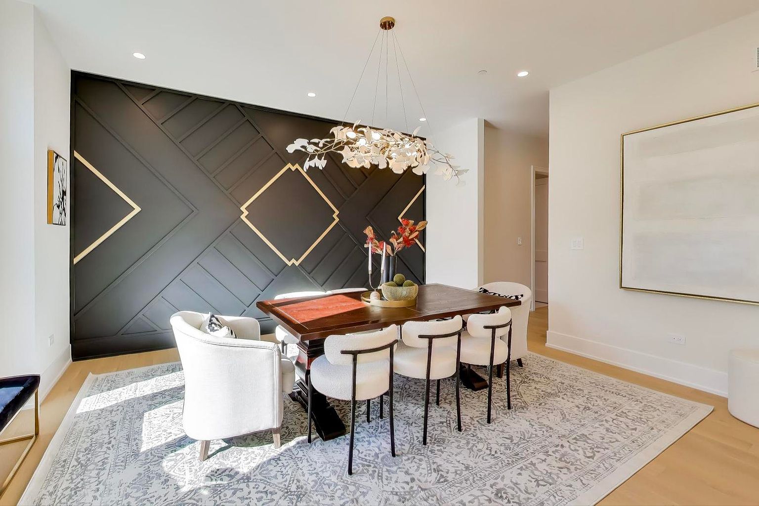 This is an interior shot of a dining room featuring a dark wood table surrounded by white chairs with black legs, set on a patterned gray rug. A modern chandelier hangs above the table, and a black accent wall with gold geometric designs adds a touch of elegance. The room is well-lit and appears spacious, creating an inviting atmosphere for dining and entertaining.