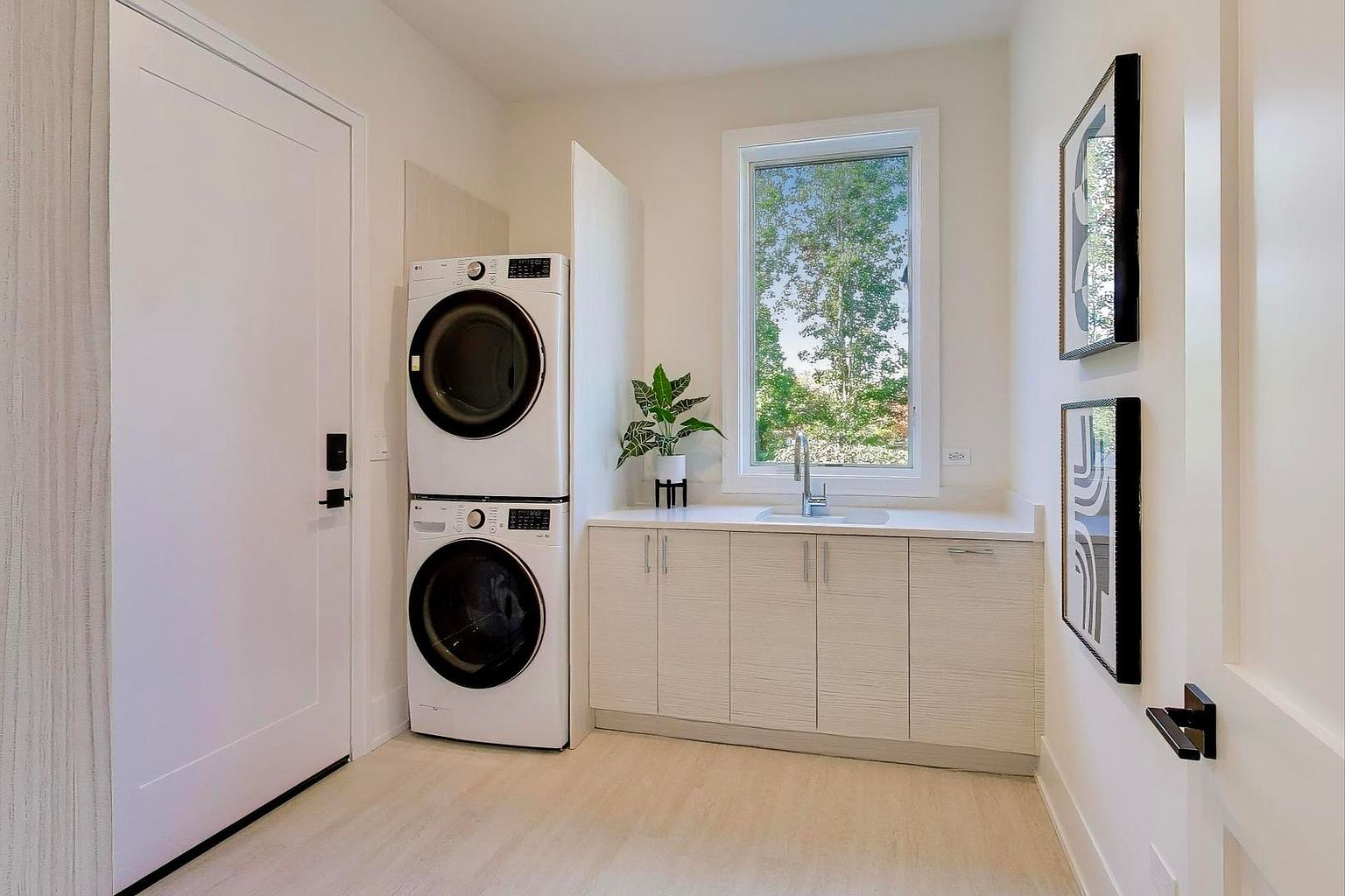 This is a well-organized laundry room featuring a stacked washer and dryer set, built-in cabinetry with a countertop, and a window providing natural light. The room has a clean and modern aesthetic with light-colored walls and flooring, complemented by minimalist artwork. The overall impression is a functional and stylish space.