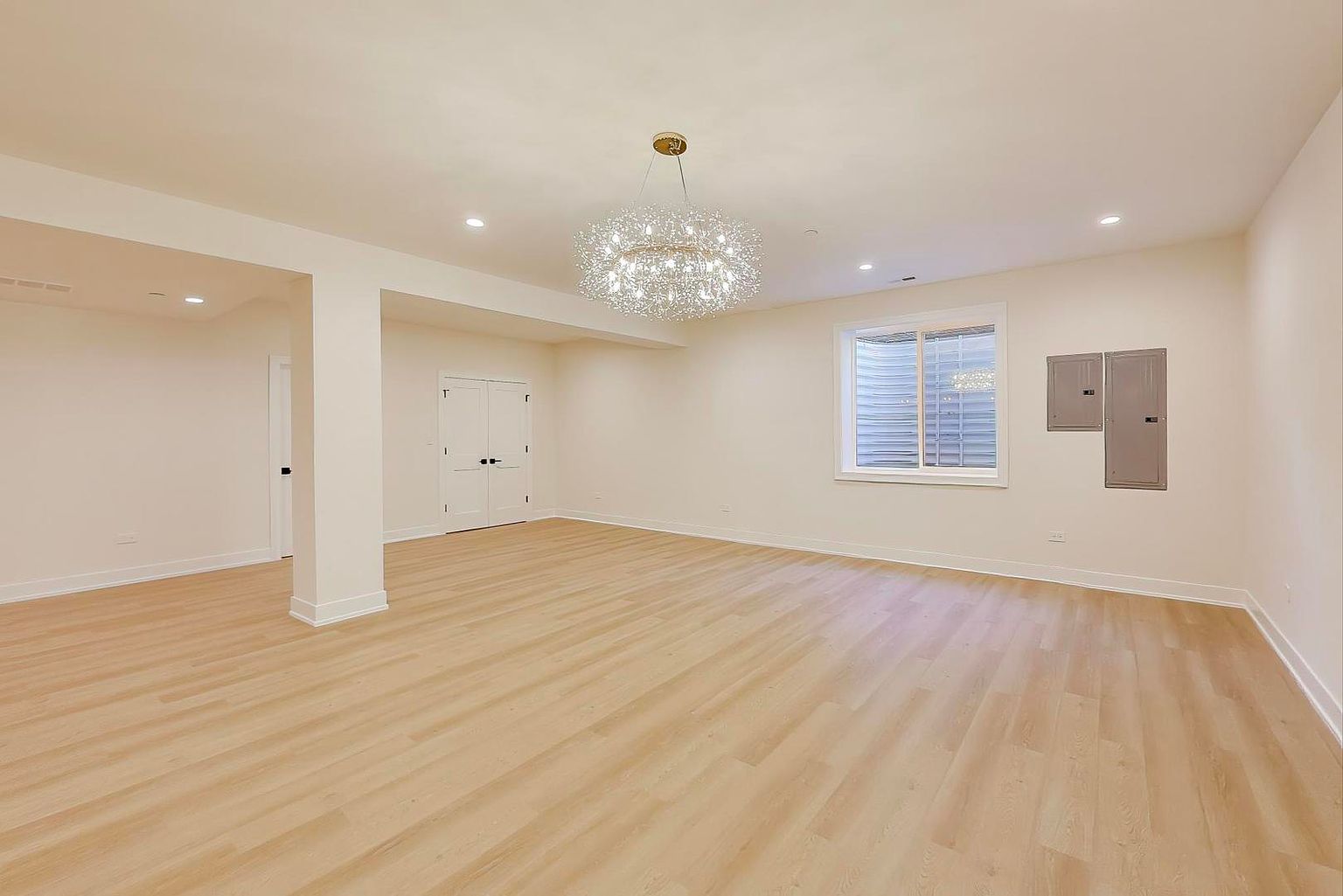 This is a spacious, finished basement featuring light wood-look flooring and neutral-colored walls. The room is well-lit with recessed lighting and a decorative chandelier, and includes a window with a window well. There are also two doors and electrical panels visible, suggesting functional areas within the basement.