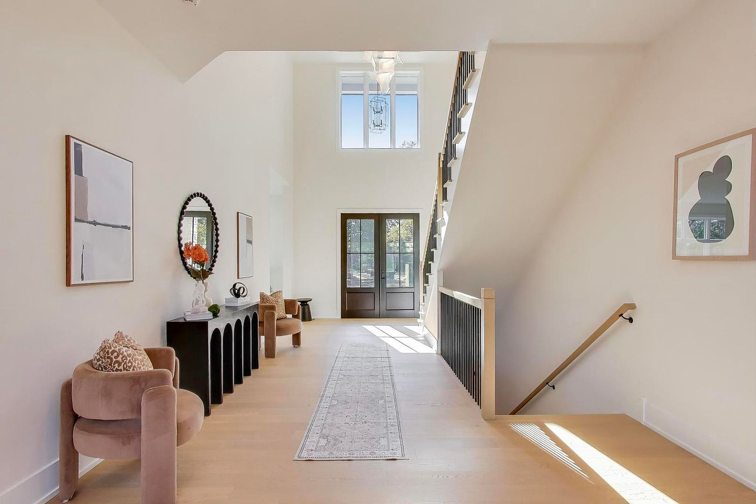This is a bright and spacious entryway featuring a modern design with clean lines and neutral colors. The foyer includes a console table with decorative items, artwork on the walls, and comfortable seating. A staircase with black railings leads to the upper level, and a long runner rug adds texture to the light wood flooring.