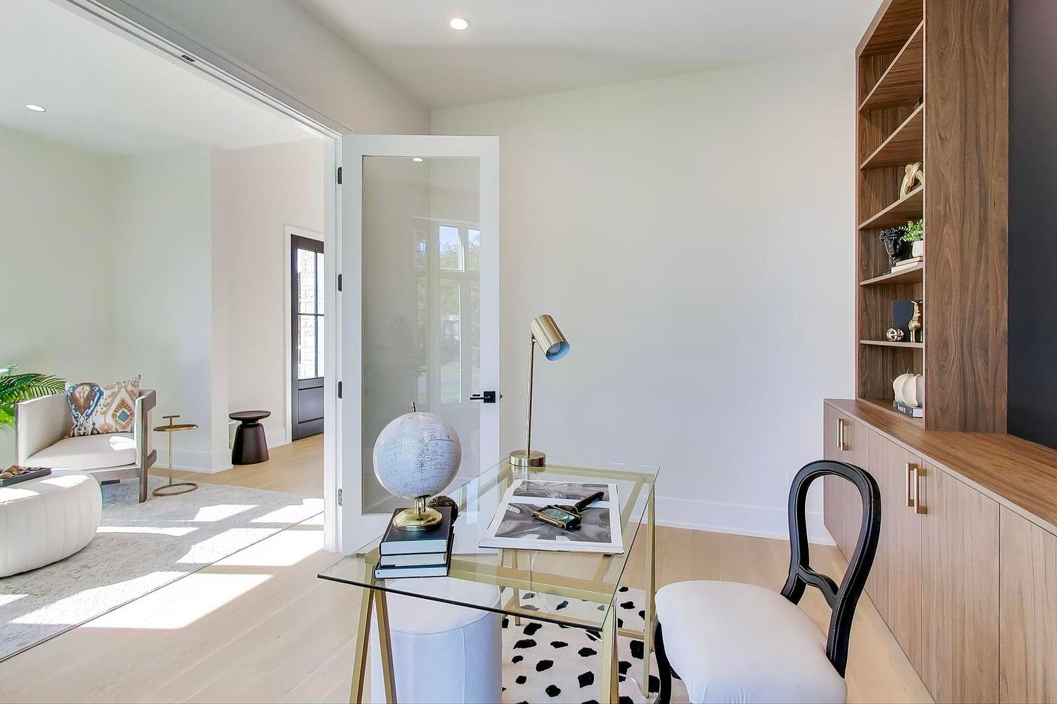 This is a bright and airy home office featuring a glass-top desk with gold accents, a globe, and a modern chair. The room has light wood floors, white walls, and a built-in bookshelf, creating a clean and sophisticated workspace. Natural light floods the room, enhancing the sense of calm and productivity.