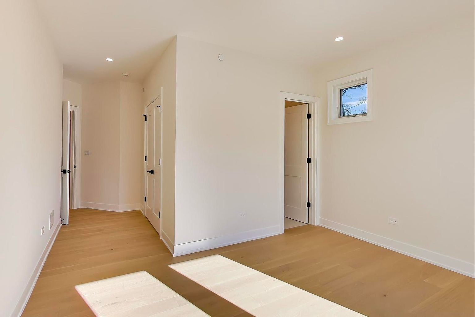 This is a bright and airy bedroom featuring light hardwood floors and white walls, creating a clean and modern aesthetic. Natural light streams in through a small window, and a doorway leads to another room, suggesting a connected layout. The room is simple yet elegant, offering a blank canvas for potential buyers.