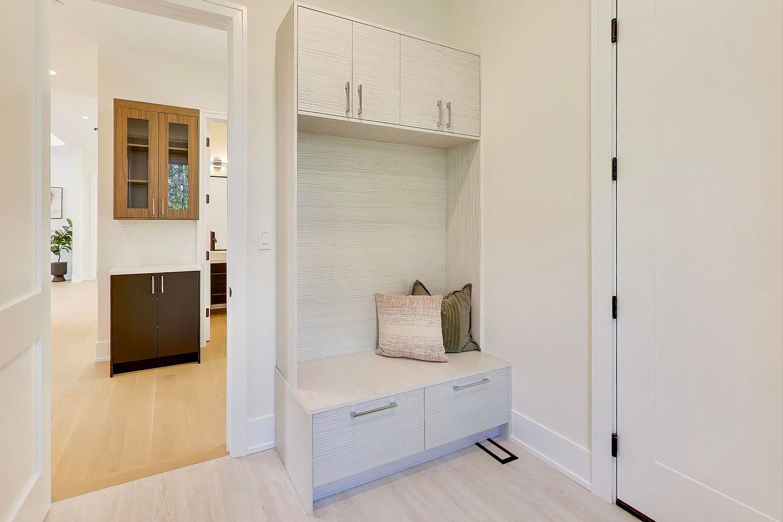 This interior shot showcases a well-lit hallway featuring a built-in storage unit with a bench seat and overhead cabinets. The color palette is neutral, with light wood tones and white walls, creating a clean and modern aesthetic. A doorway leads to another room, providing a glimpse of additional cabinetry and hardwood flooring.