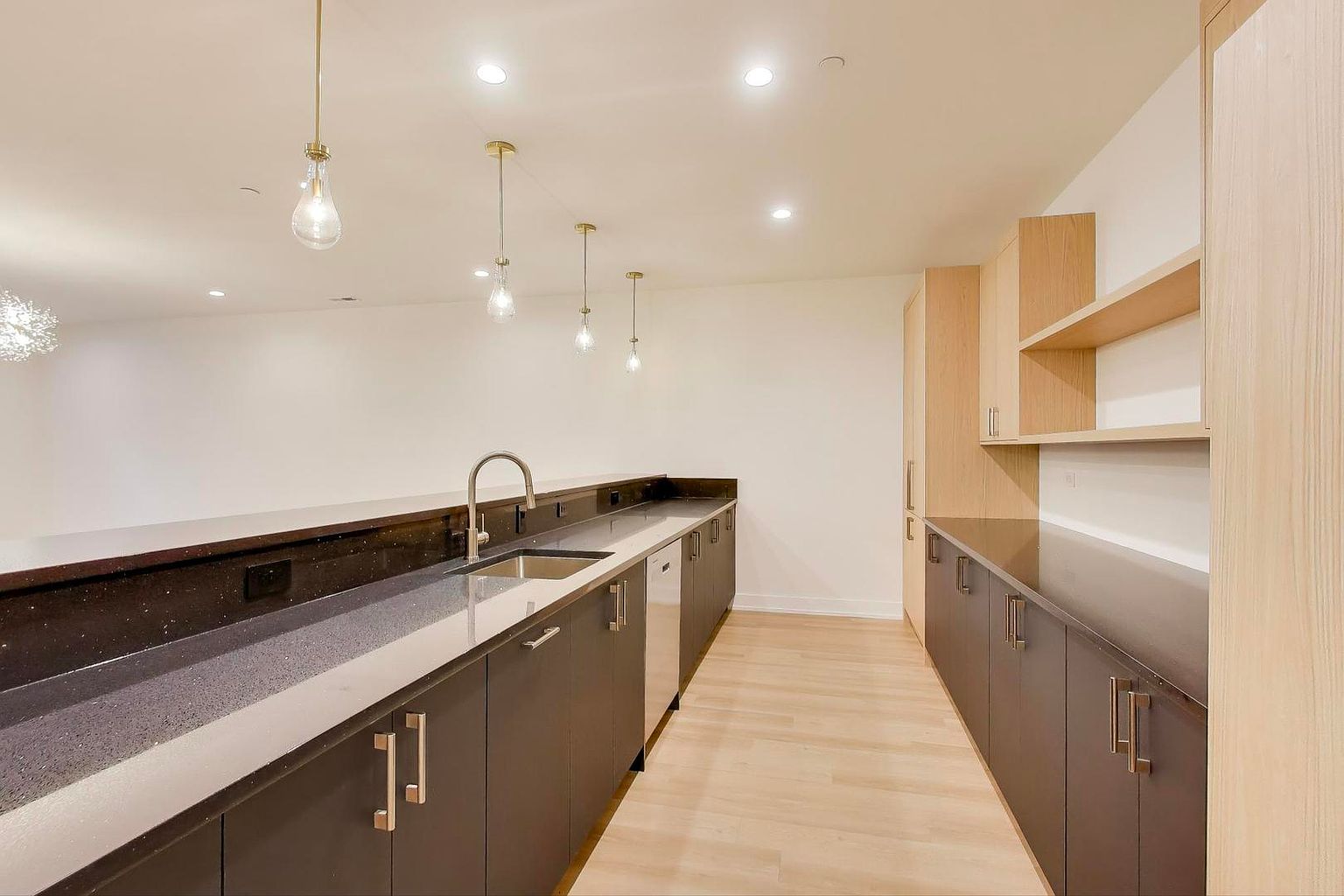 This is a modern kitchen featuring sleek, dark cabinetry with minimalist hardware and light wood accents. The countertops are a dark, speckled material, and the flooring is light-colored wood. Pendant lights hang above the sink area, and the overall impression is clean and contemporary.