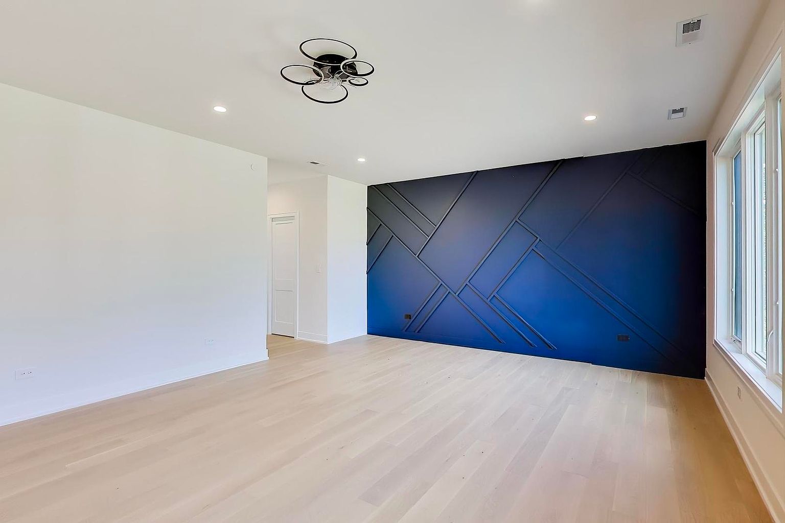 This is an interior shot of a modern living room featuring light wood flooring and a striking dark blue accent wall with geometric trim. The room is well-lit with recessed lighting and natural light from a window on the right. A contemporary ceiling light fixture adds a stylish touch to the space, creating a clean and inviting atmosphere.