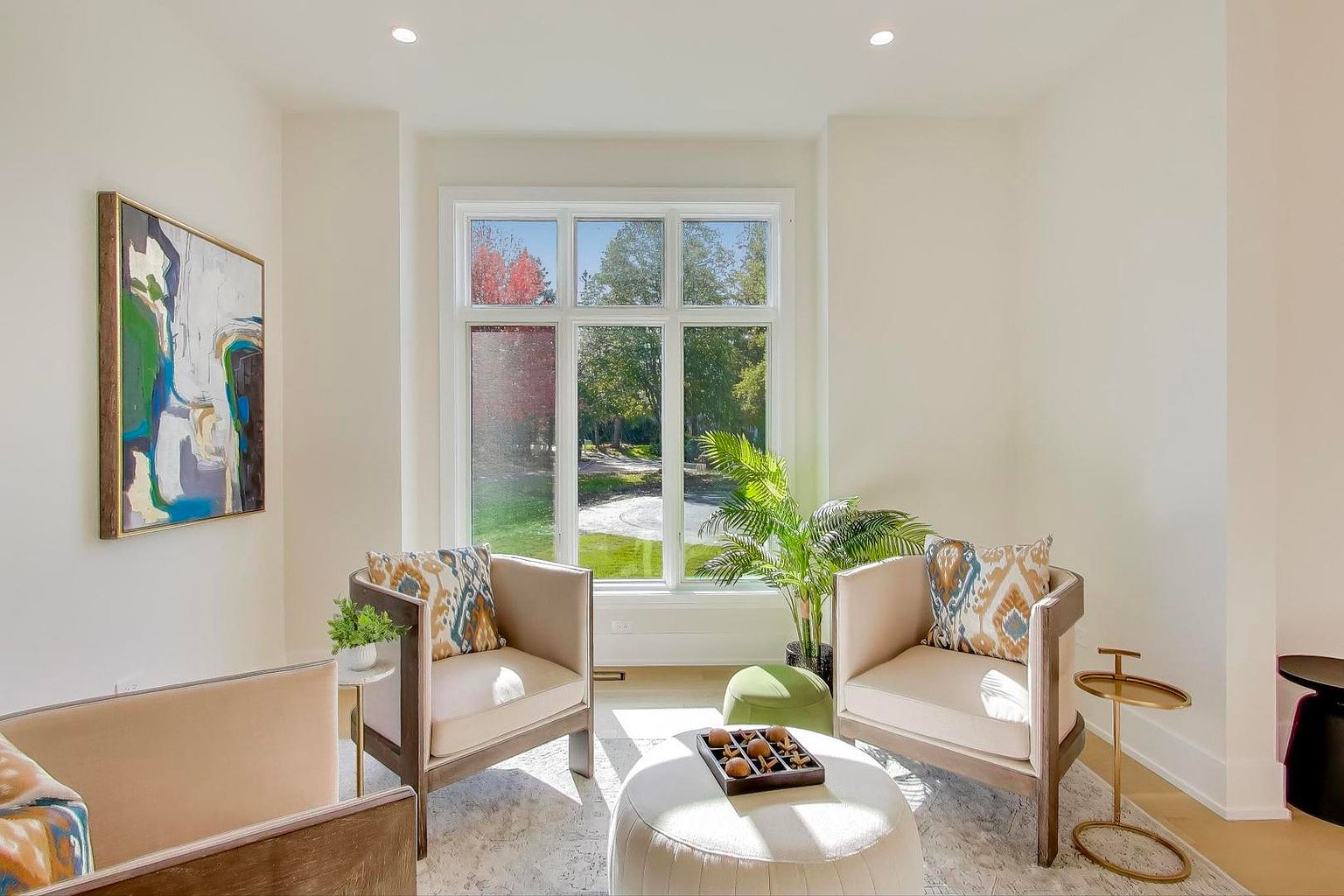 This is an interior shot of a bright and airy living room. The room features two armchairs with patterned pillows, a white ottoman with a tray of items, and a large window offering a view of a green landscape. An abstract painting hangs on the wall, and the overall style is modern and inviting.