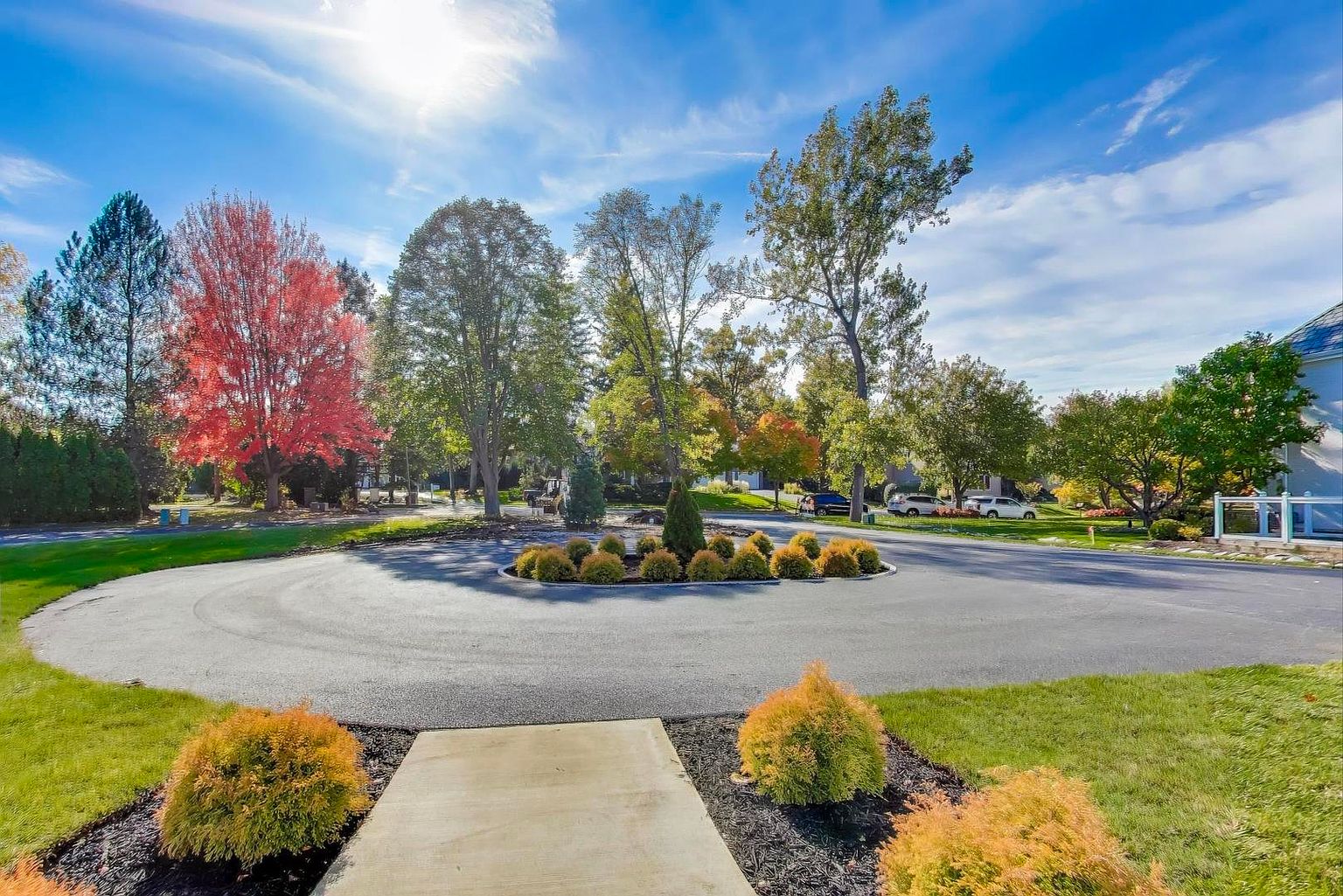 This is a front view of a property featuring a circular driveway with a landscaped island in the center. The property is surrounded by mature trees, some displaying vibrant fall colors, under a bright blue sky with scattered clouds. A concrete walkway leads to the property, flanked by well-maintained lawns and shrubbery, creating a welcoming and picturesque curb appeal.