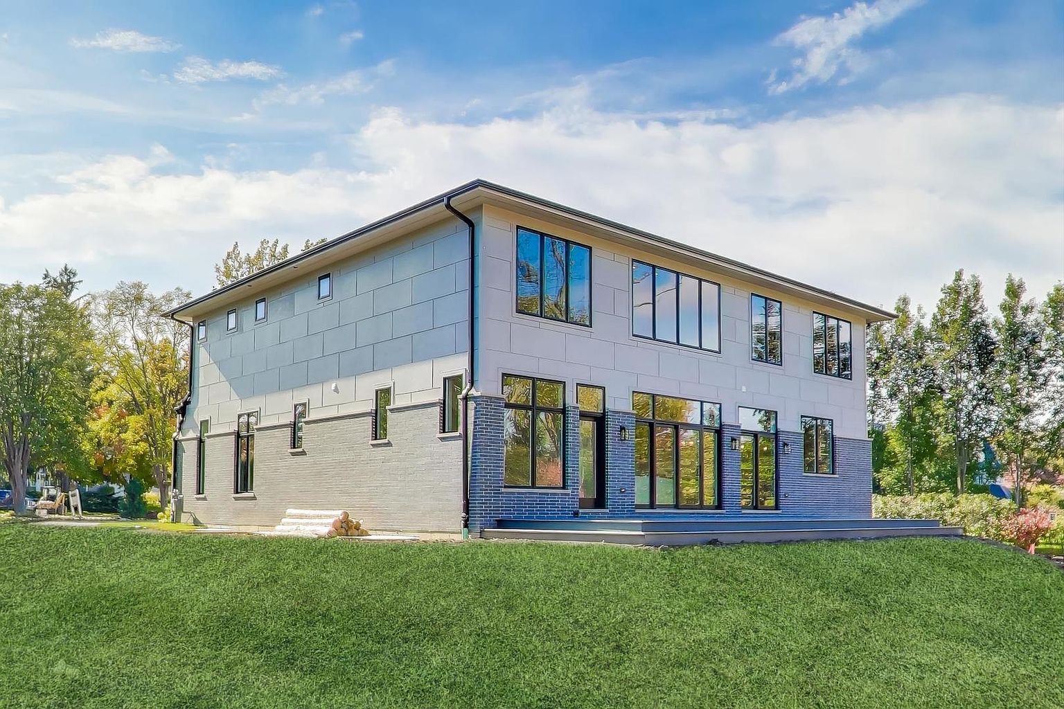 This is a rear view of a modern two-story house featuring a combination of gray brick and panel siding. Large windows and sliding glass doors provide ample natural light and access to an outdoor deck area. The well-maintained lawn and surrounding trees enhance the property's appeal.