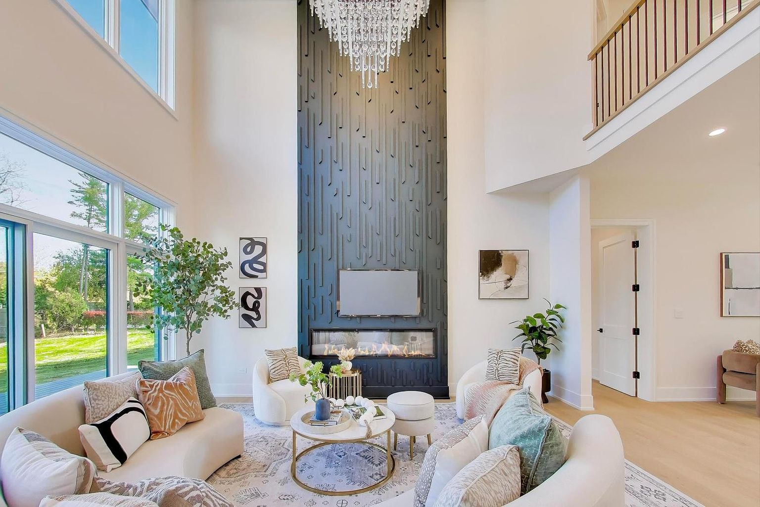 This is a bright and airy living room featuring a soaring ceiling and a modern fireplace with a television mounted above. The room is furnished with comfortable, curved sofas and armchairs arranged around a round coffee table, creating an inviting and stylish space. Large windows provide ample natural light and views of the outdoors.