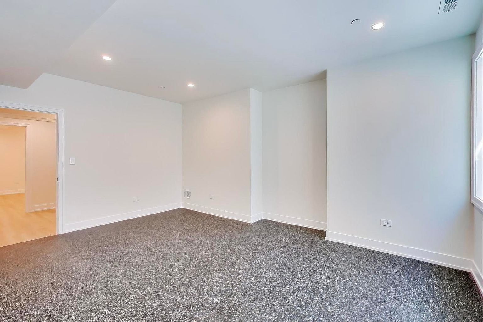 The image showcases a well-lit, empty bedroom with neutral gray carpeting and white walls, creating a blank canvas for potential buyers. Recessed lighting provides ample illumination, and a doorway hints at an en-suite bathroom or walk-in closet. The room's clean lines and minimalist design offer a sense of spaciousness and versatility.