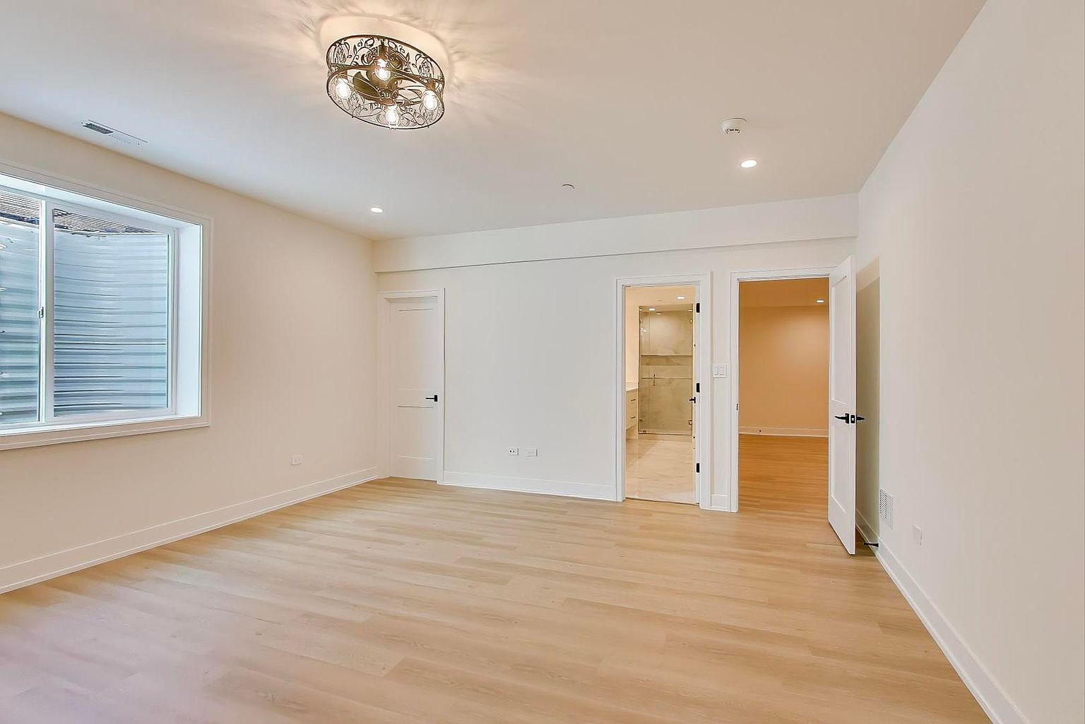 The image showcases a bright and airy bedroom with light wood flooring and neutral-colored walls. A window provides natural light, and a decorative ceiling light fixture adds a touch of elegance. Two doorways lead to other rooms, suggesting an en-suite bathroom and possibly a hallway or closet.