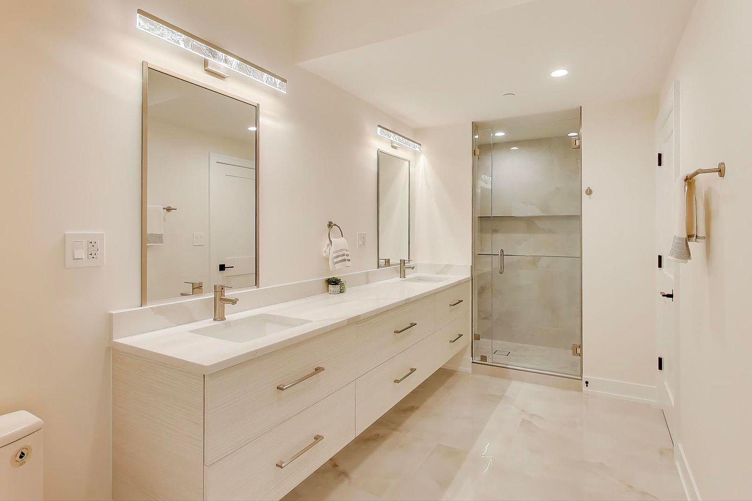 This is a bright and modern primary bathroom featuring a double vanity with a white countertop and light wood-grain cabinets. Two large mirrors are illuminated by contemporary light fixtures. A glass-enclosed shower is visible in the background, and the floor is covered in light-colored tile, creating a clean and luxurious atmosphere.