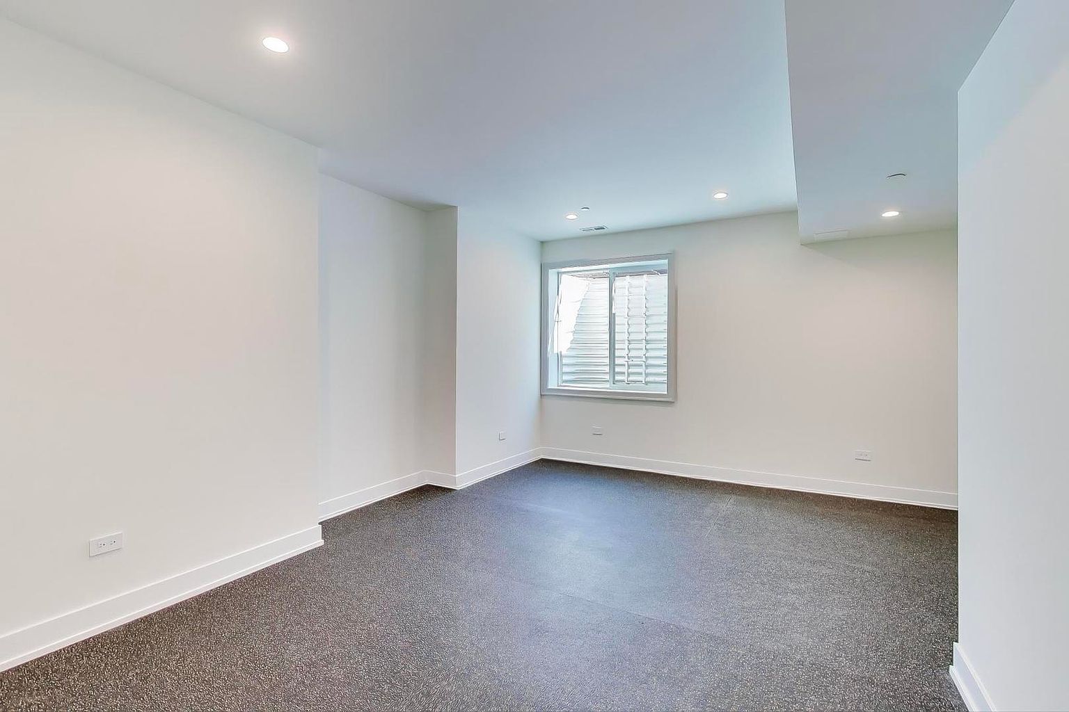 The image showcases a clean, unfinished basement with a neutral color palette. The walls are painted white, and the flooring appears to be a dark gray or black. A window provides natural light, and recessed lighting illuminates the space, creating a bright and open feel.