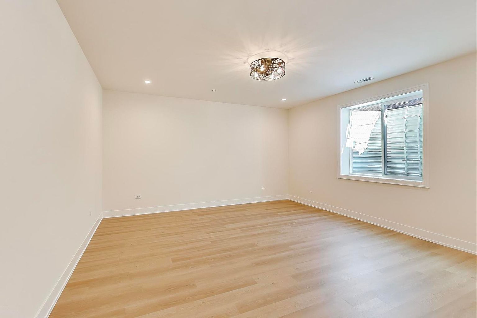 This is an interior shot of a bedroom, likely a guest or secondary bedroom, featuring light wood flooring and neutral-colored walls. A window provides natural light, and a decorative ceiling light fixture adds a touch of elegance. The room is empty, showcasing its spaciousness and potential for customization.