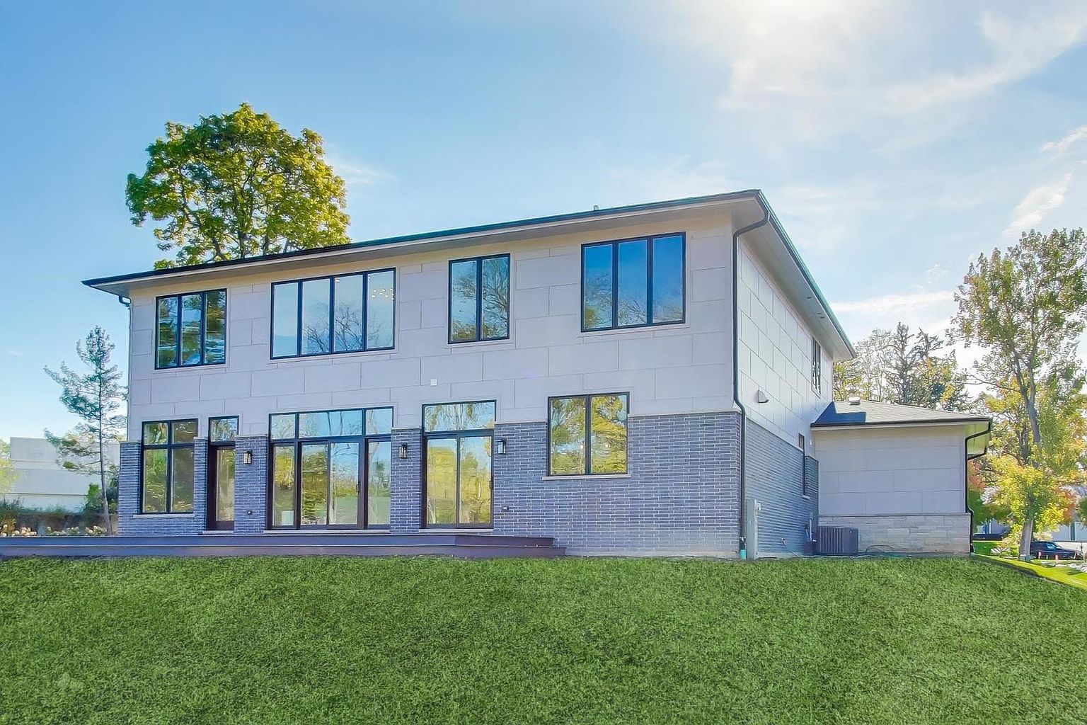This is a rear view of a modern two-story home featuring a combination of light gray paneling and gray brick on the lower level. Large windows with black frames provide ample natural light, and a well-maintained green lawn extends from the house. The overall impression is clean, contemporary, and inviting.