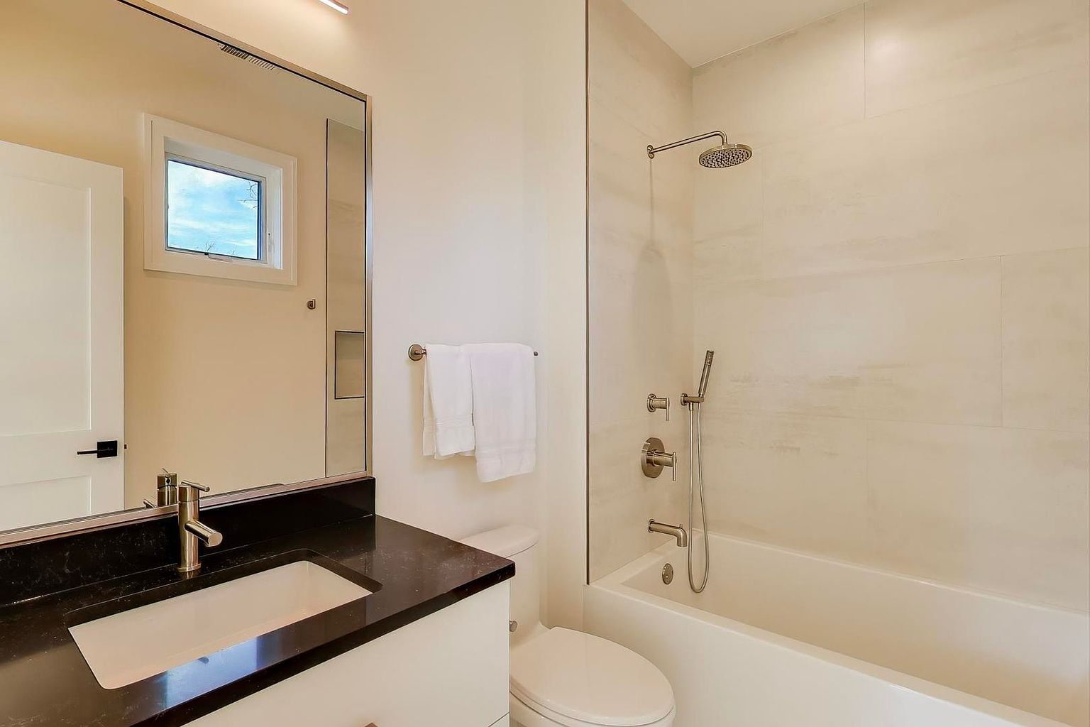 This is a well-lit bathroom featuring a modern design with a black countertop and white cabinets. A large mirror reflects the space, including a window. The shower/tub area is tiled in a light color, and the fixtures are brushed nickel, creating a clean and contemporary look.
