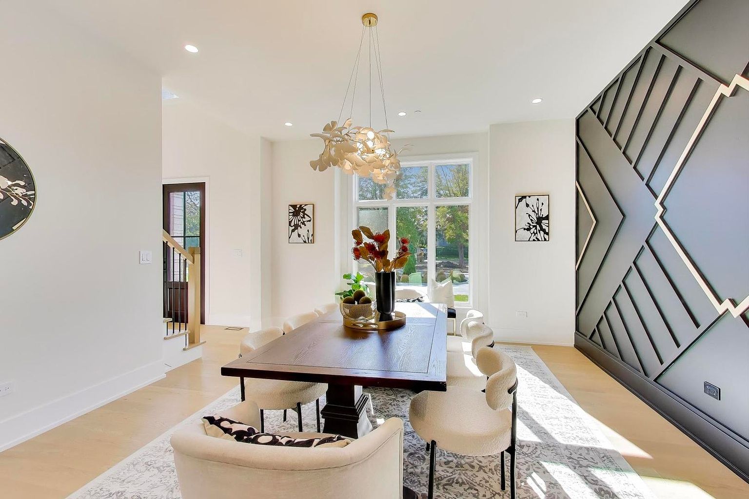 This is an interior shot of a dining room featuring a dark wood dining table surrounded by cream-colored chairs. A modern chandelier hangs above the table, and a large window provides natural light. The room is decorated with artwork and a unique geometric wall design, creating a sophisticated and inviting atmosphere.