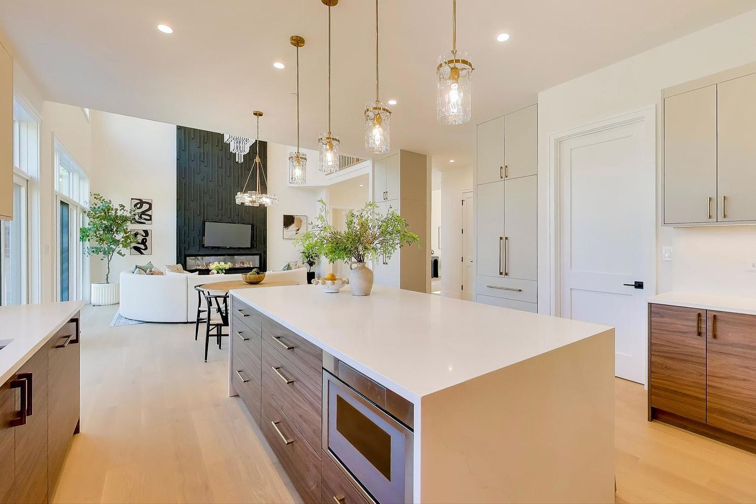 This is a bright and open kitchen and living area featuring a large kitchen island with a white countertop and wood-paneled cabinets. The kitchen includes modern appliances and pendant lighting, while the living area has a fireplace and comfortable seating. The overall design is contemporary and inviting, perfect for entertaining.