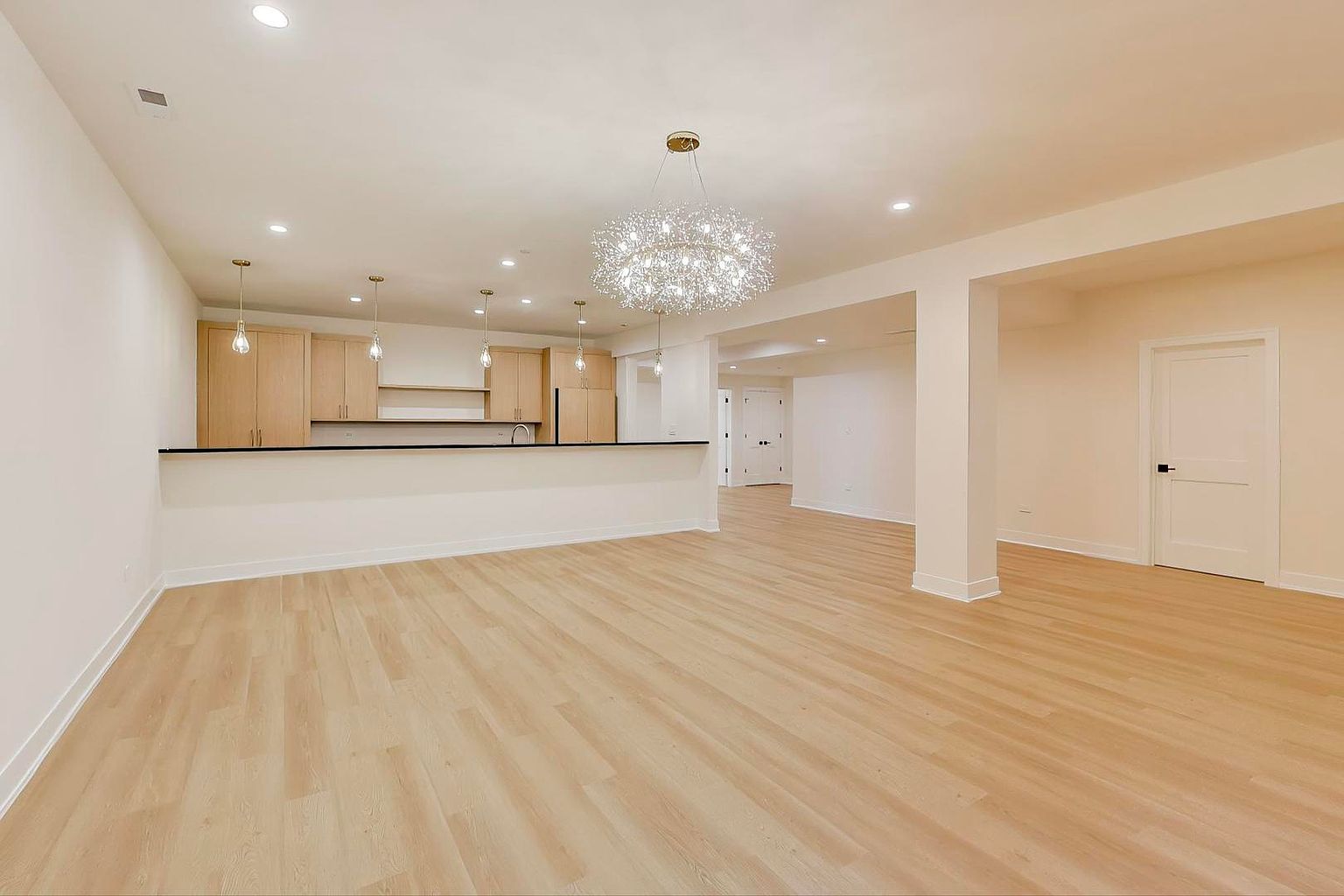 This is a bright and spacious living room with light hardwood floors and neutral-colored walls. The open floor plan seamlessly connects to the kitchen area, featuring modern cabinetry and a breakfast bar. A stylish chandelier serves as a focal point, enhancing the contemporary aesthetic of the space.