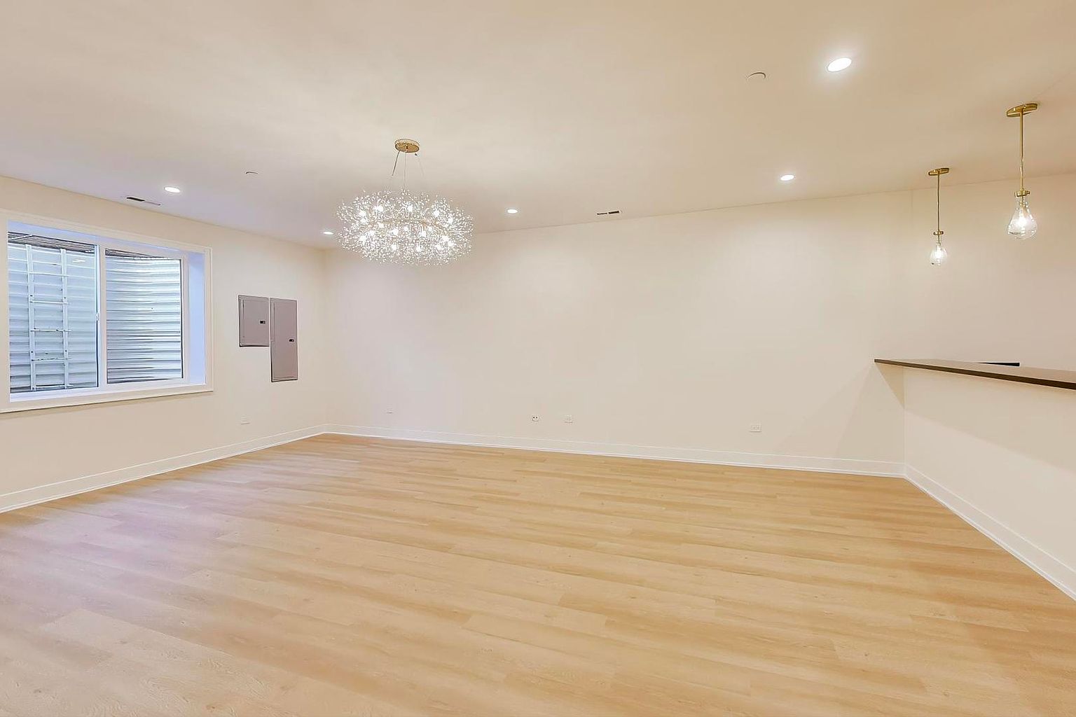 This is a spacious, well-lit basement featuring light wood flooring and white walls. A modern chandelier hangs from the ceiling, and there's a window providing natural light. A built-in bar area is visible on the right side of the room, suggesting potential for entertainment or relaxation.