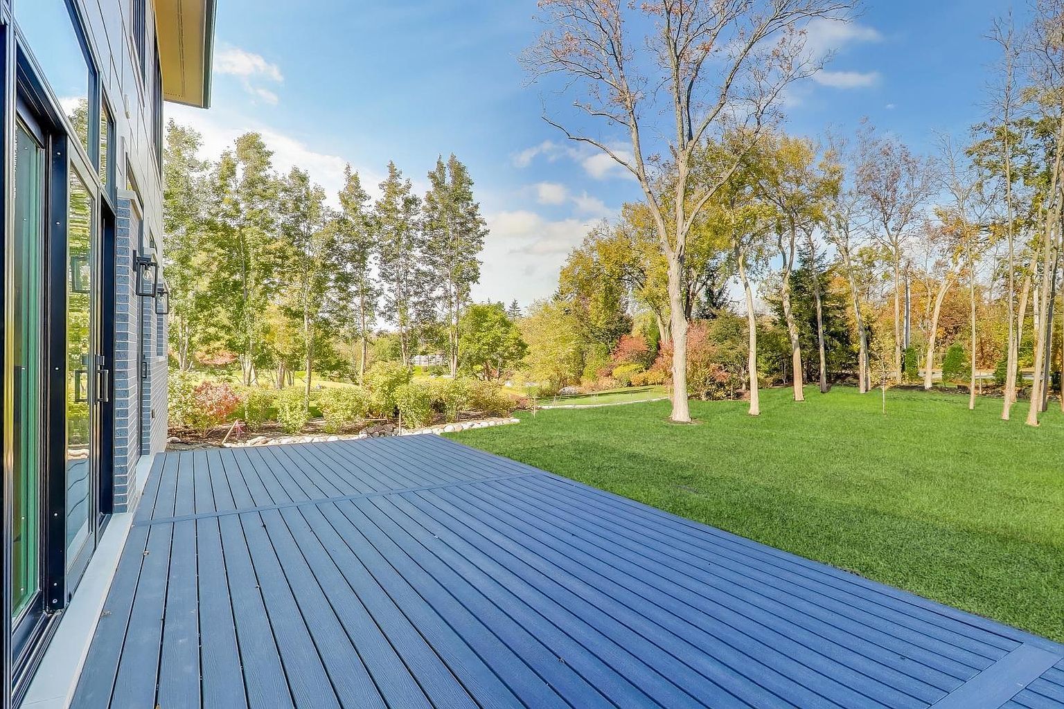 This image showcases a spacious blue deck extending from a modern home into a lush green yard. The deck's clean lines and expansive size suggest an ideal space for outdoor entertaining. Beyond the deck, the well-maintained lawn is bordered by mature trees and landscaping, creating a serene and private backyard setting.