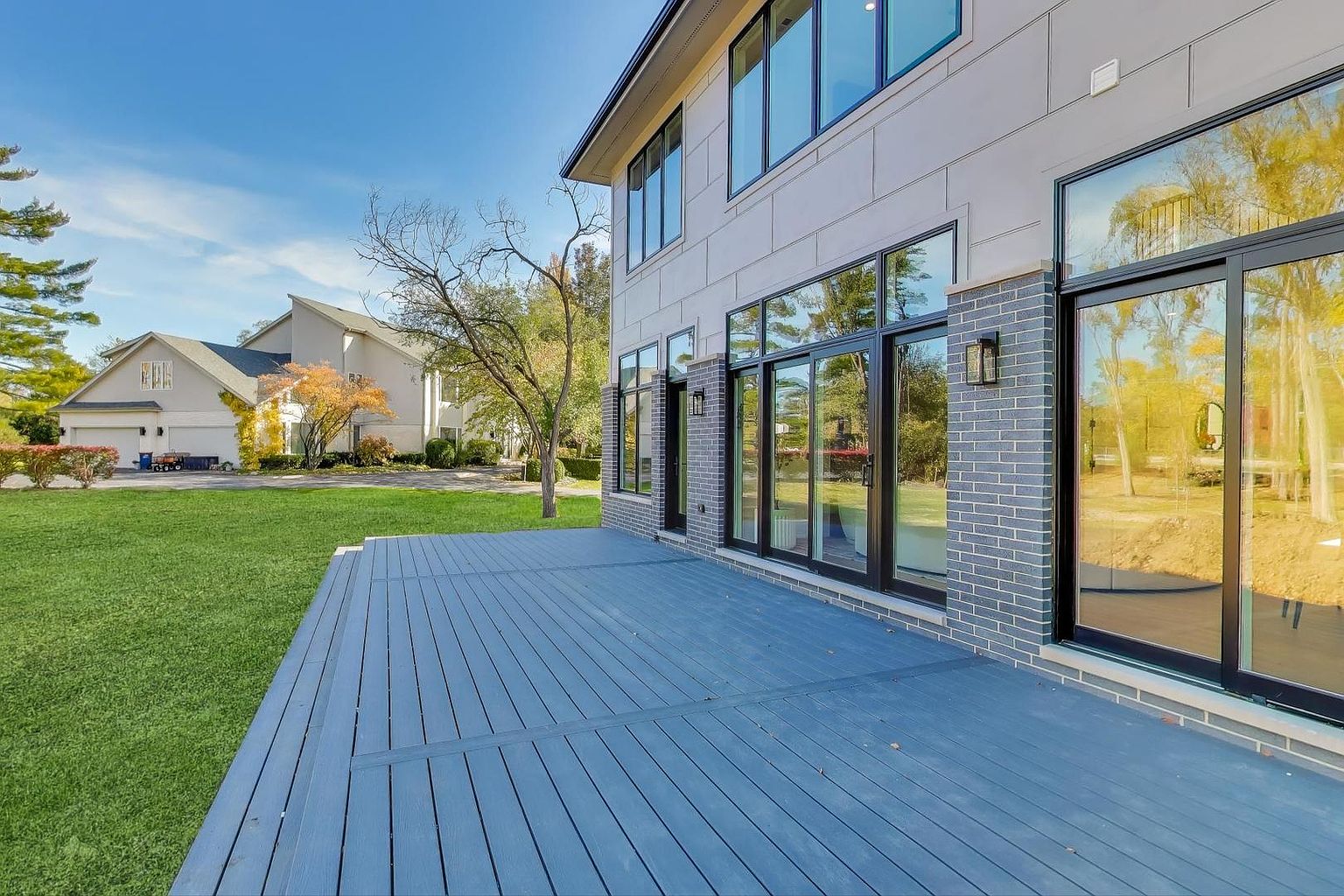 This image showcases a modern home's exterior, focusing on a spacious blue-toned deck. The deck is constructed with parallel planks and provides access to the home through large, black-framed sliding glass doors, which are flanked by brick pillars. The view includes a well-maintained lawn and a glimpse of neighboring houses under a clear sky, creating an inviting outdoor living space.