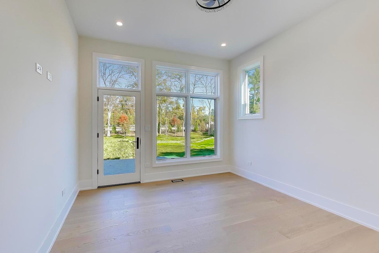 This is a bright and airy sunroom featuring light hardwood floors, white walls, and large windows that offer a view of the outdoors. The room is well-lit with natural light, creating a welcoming and serene atmosphere. The door leads to the outside, enhancing the indoor-outdoor flow.