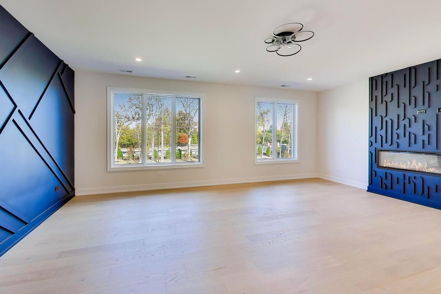 This is an interior shot of a modern living room featuring light hardwood floors and white walls. A striking blue accent wall with geometric paneling is on the left, while a modern fireplace with a blue decorative surround is on the right. Large windows provide natural light, and a contemporary ceiling light fixture adds a stylish touch.