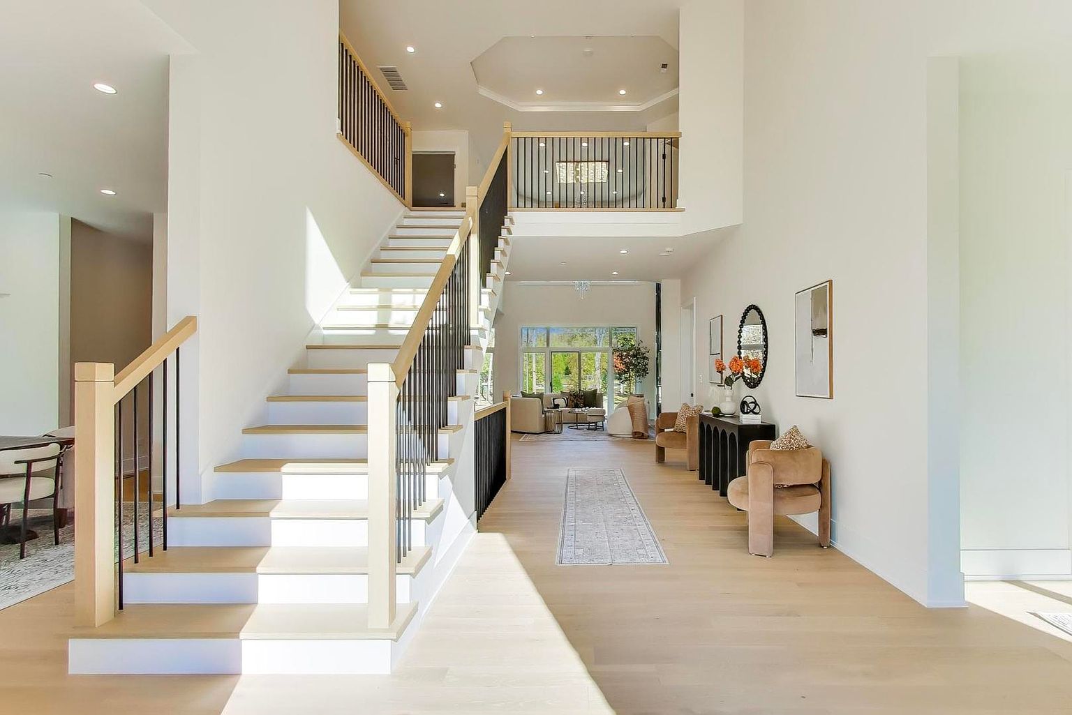 This is an interior shot of a grand foyer featuring a staircase with light wood treads and white risers, complemented by a black metal and light wood railing. The space is bright and airy, with light hardwood flooring and white walls, leading to a living area visible in the background. The overall impression is one of spaciousness and modern elegance.