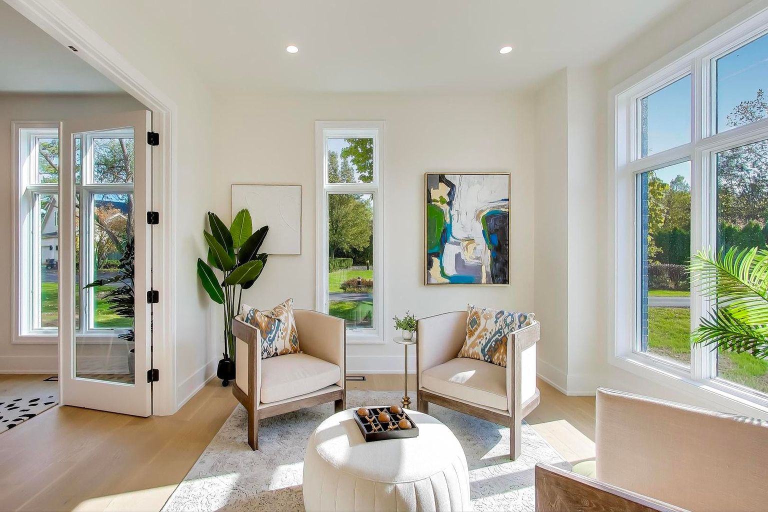 This is a bright and airy living room featuring two armchairs with patterned pillows, a round ottoman, and abstract artwork. Large windows provide ample natural light and views of the outdoors. The room is decorated in neutral tones with wood accents, creating a cozy and inviting atmosphere.
